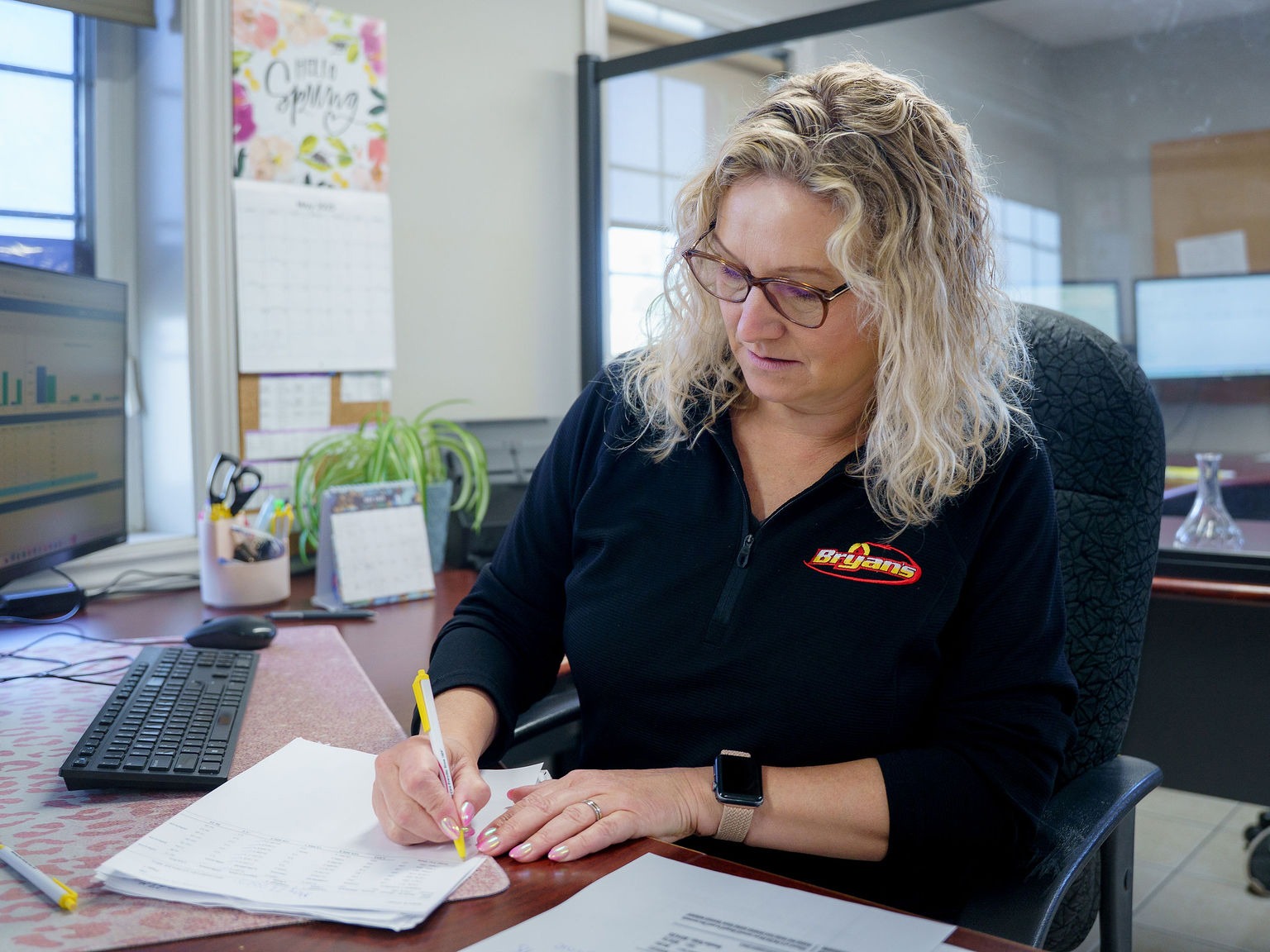 A person with glasses is working at a desk, writing on papers. Computer monitors and office supplies surround them. Calendar reads "Hello Spring."