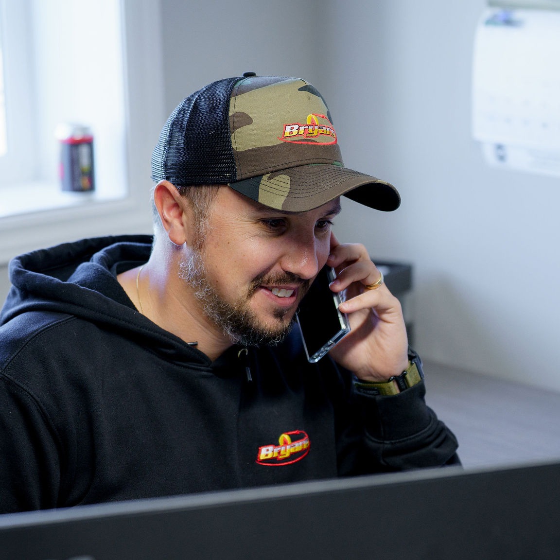 A person in a cap and sweatshirt talks on the phone at a desk. A can and calendar are visible in the background.