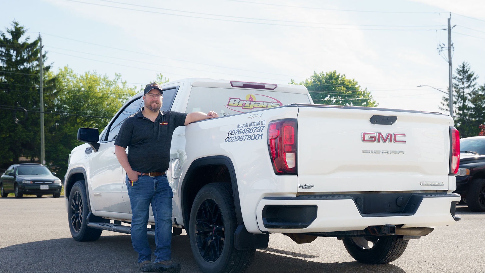 A person wearing a cap and glasses stands beside a white GMC Sierra truck in a parking lot with trees in the background.
