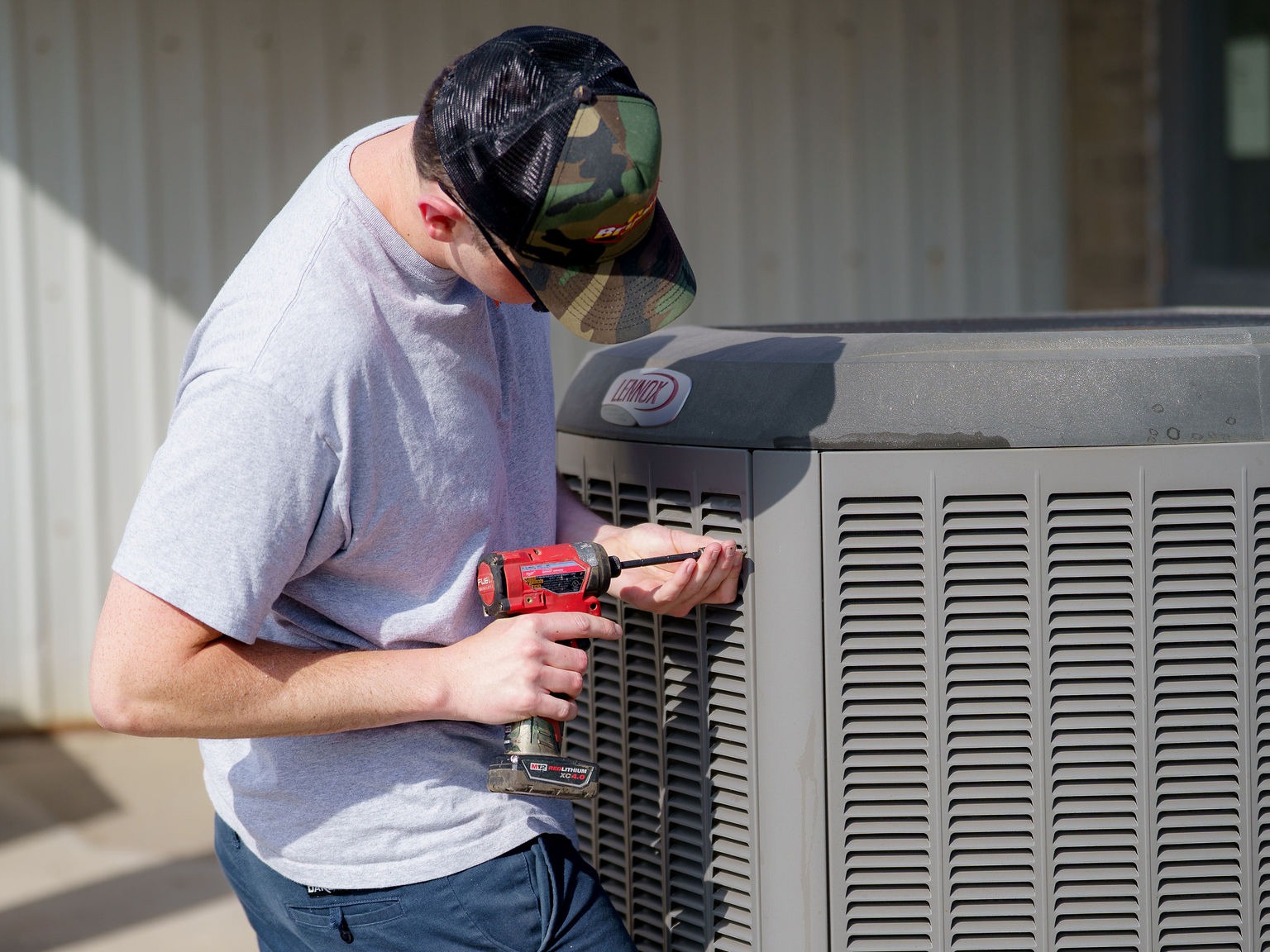 A person wearing a cap uses a red drill to work on an air conditioning unit outside, beside a corrugated metal wall.