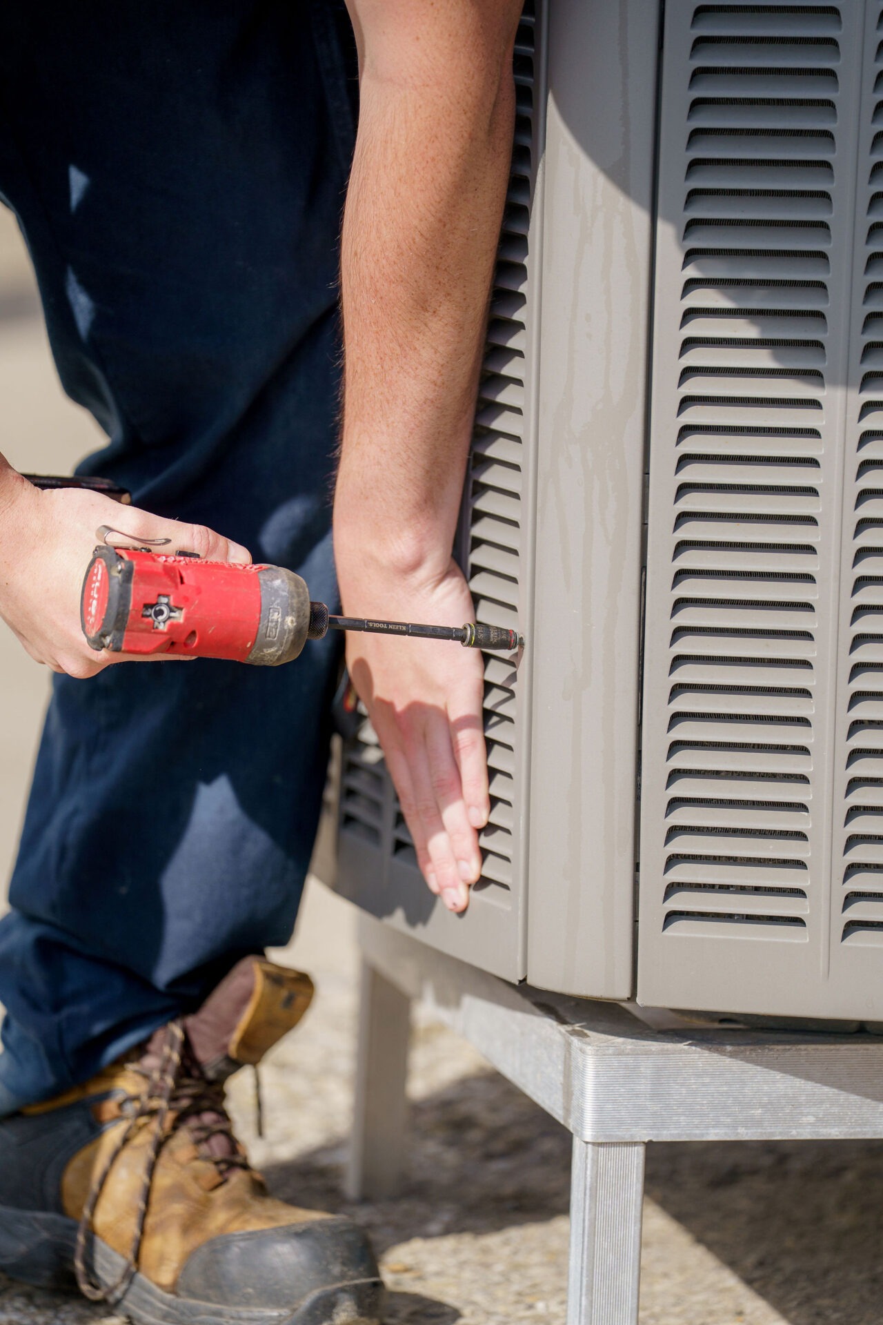 A person uses a red drill to repair or assemble a metal ventilation unit, wearing work boots and dark pants.