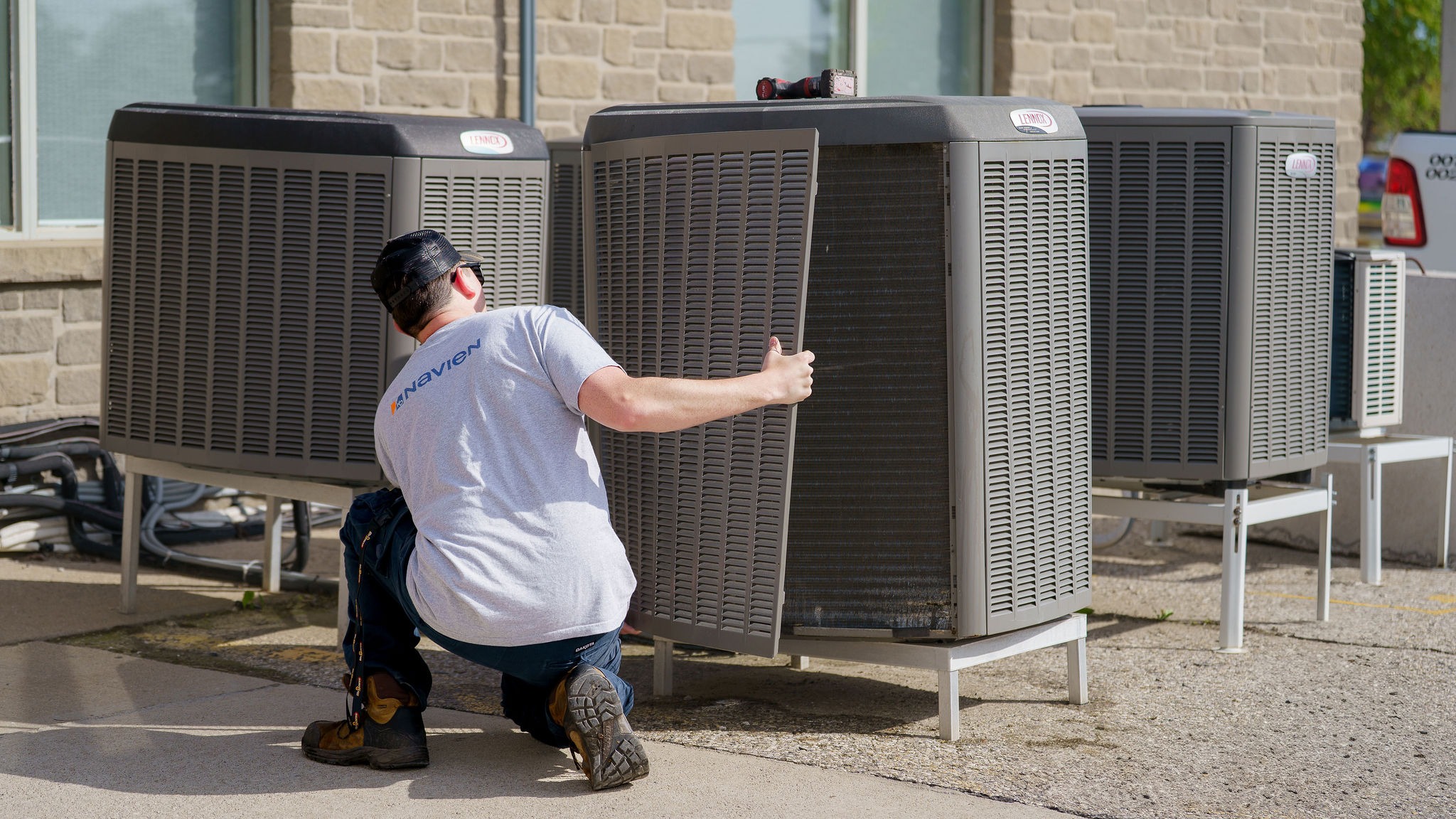 A person in a gray shirt kneels, inspecting a large outdoor air conditioning unit, near a brick building on a sunny day.