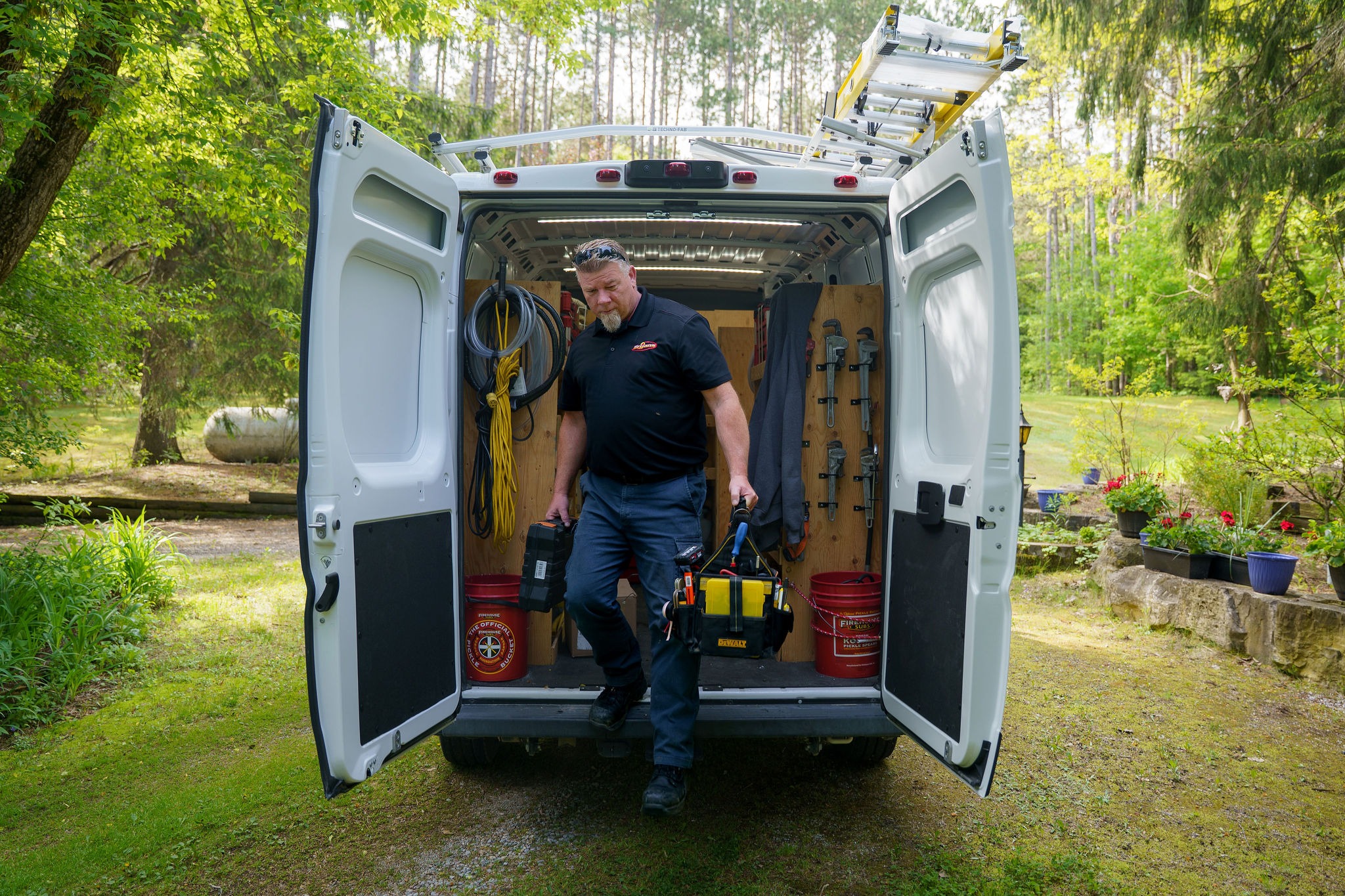 A person with tools exits a white van parked in a lush, green, wooded area with potted plants nearby.