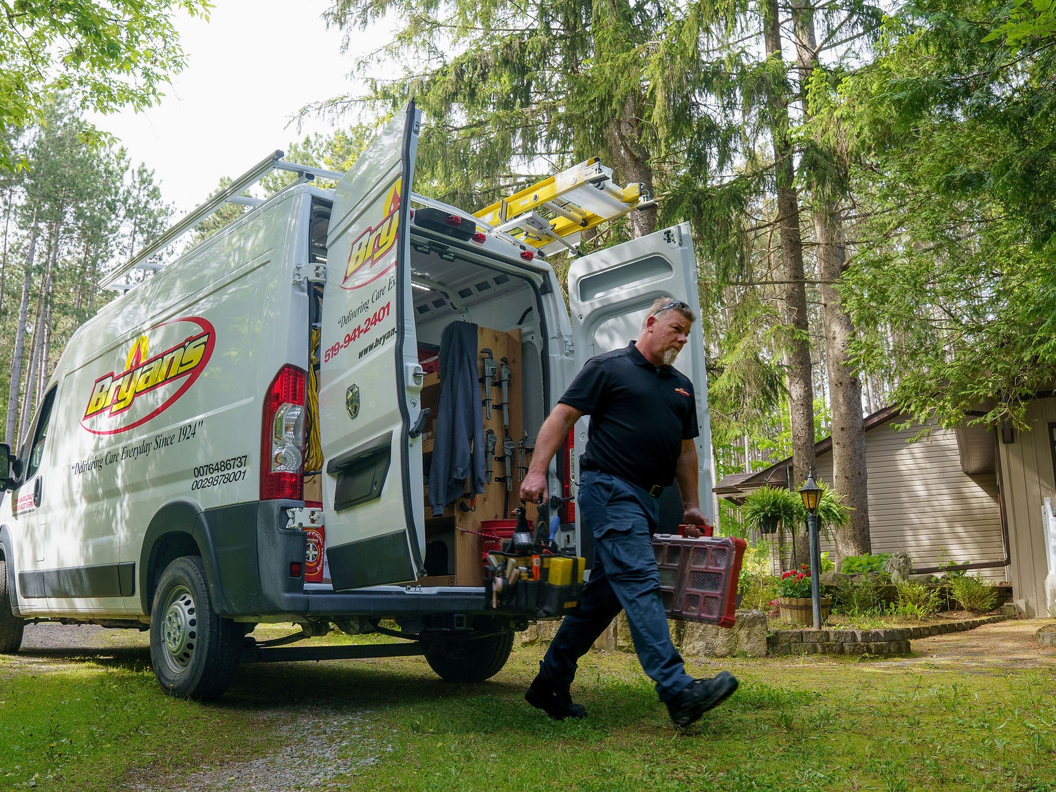 A person carries tools from a Bryans van parked near a residential property with surrounding trees and greenery.
