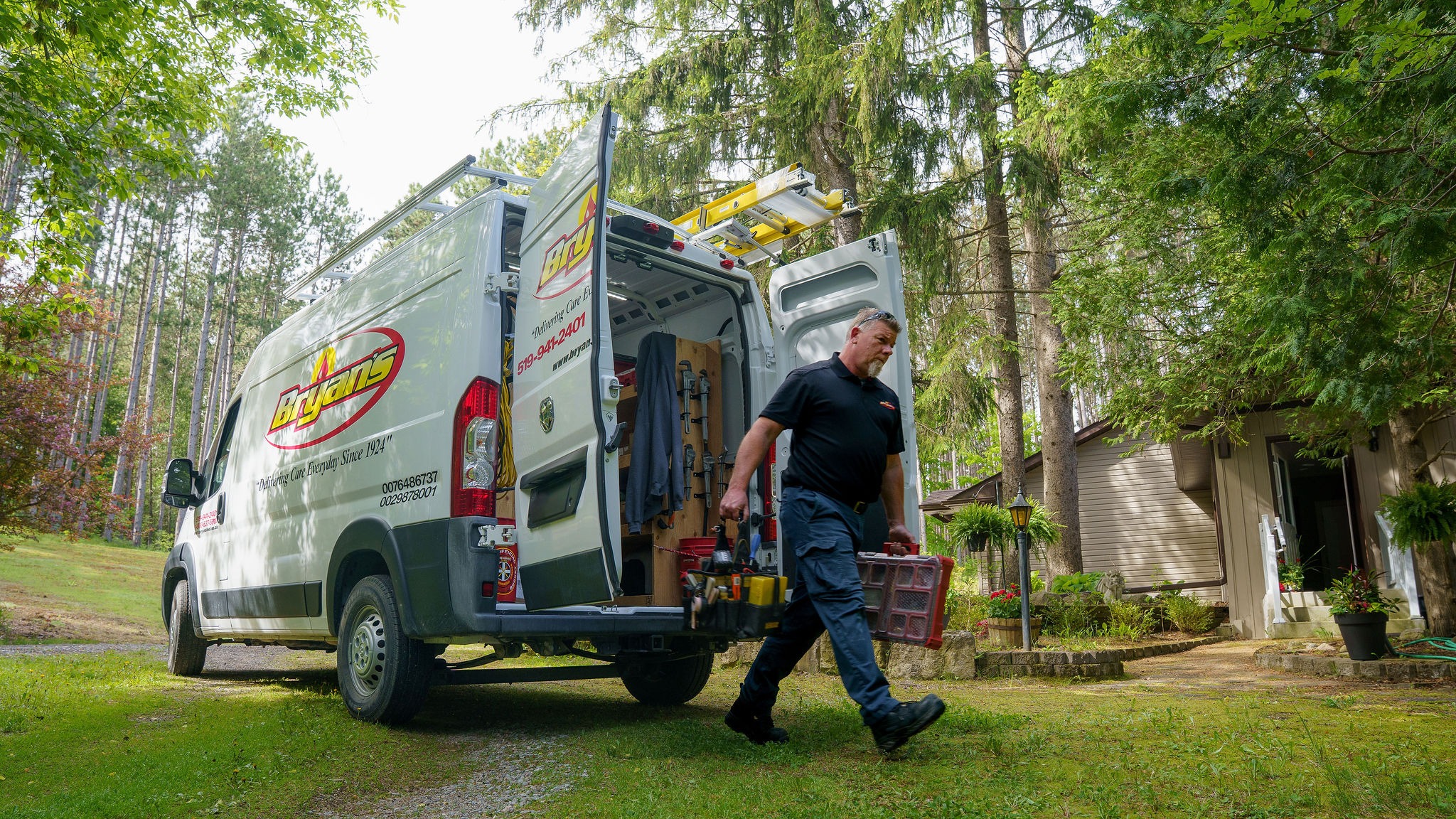 A person carries tools from a Bryans vehicle in a lush, wooded yard approaching a house surrounded by tall trees and greenery.