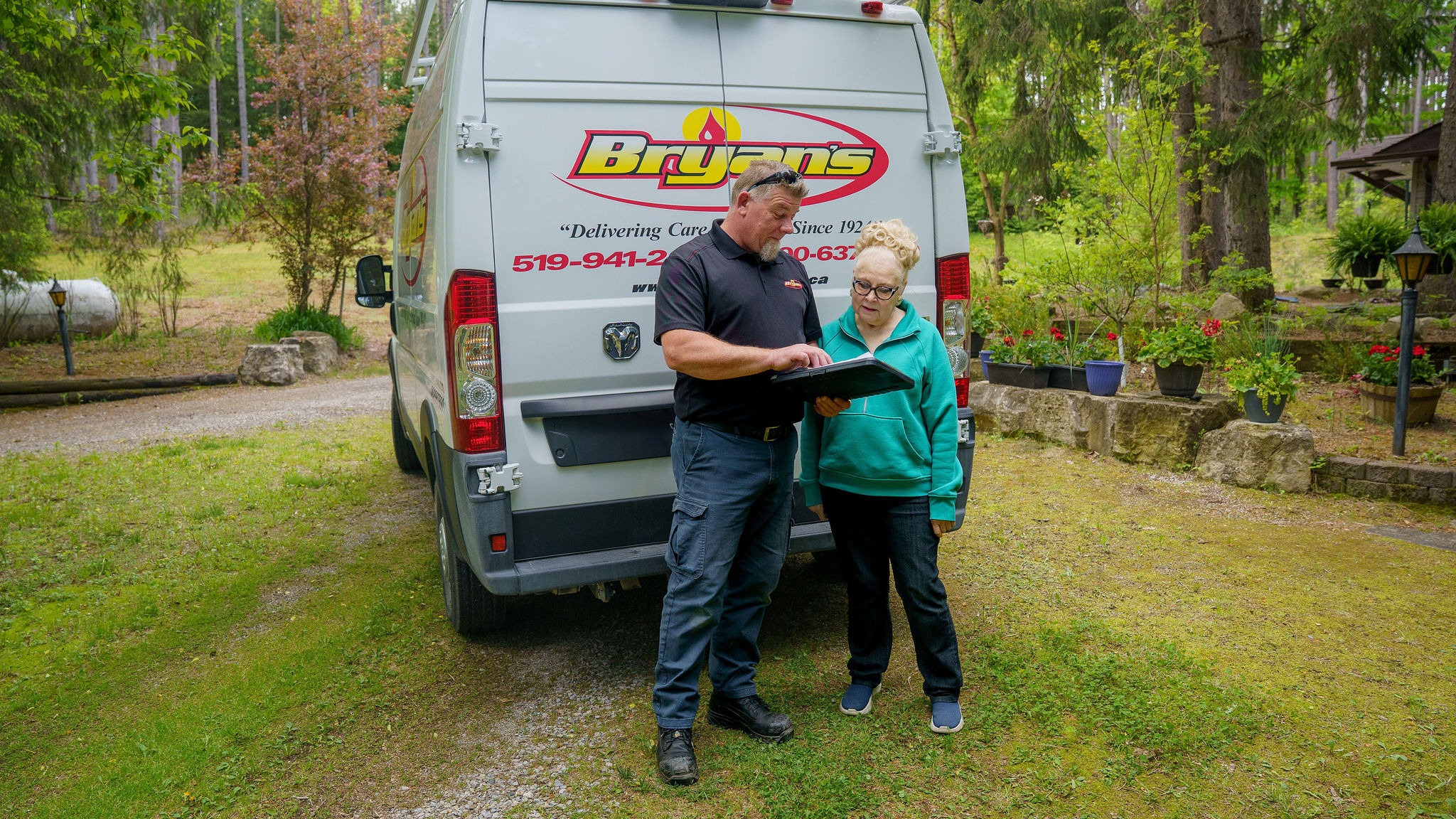 Two people stand by a parked van, reviewing a document. The setting is a lush garden area with potted plants and trees.