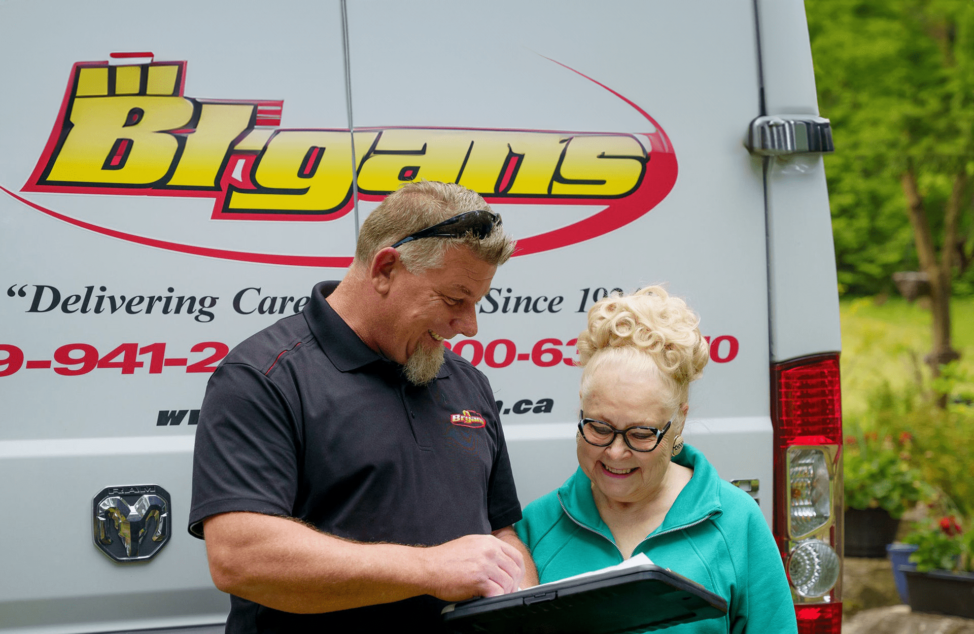 Two people smiling while looking at a document, standing beside a Bigam's van with visible contact information and logo in an outdoor setting.