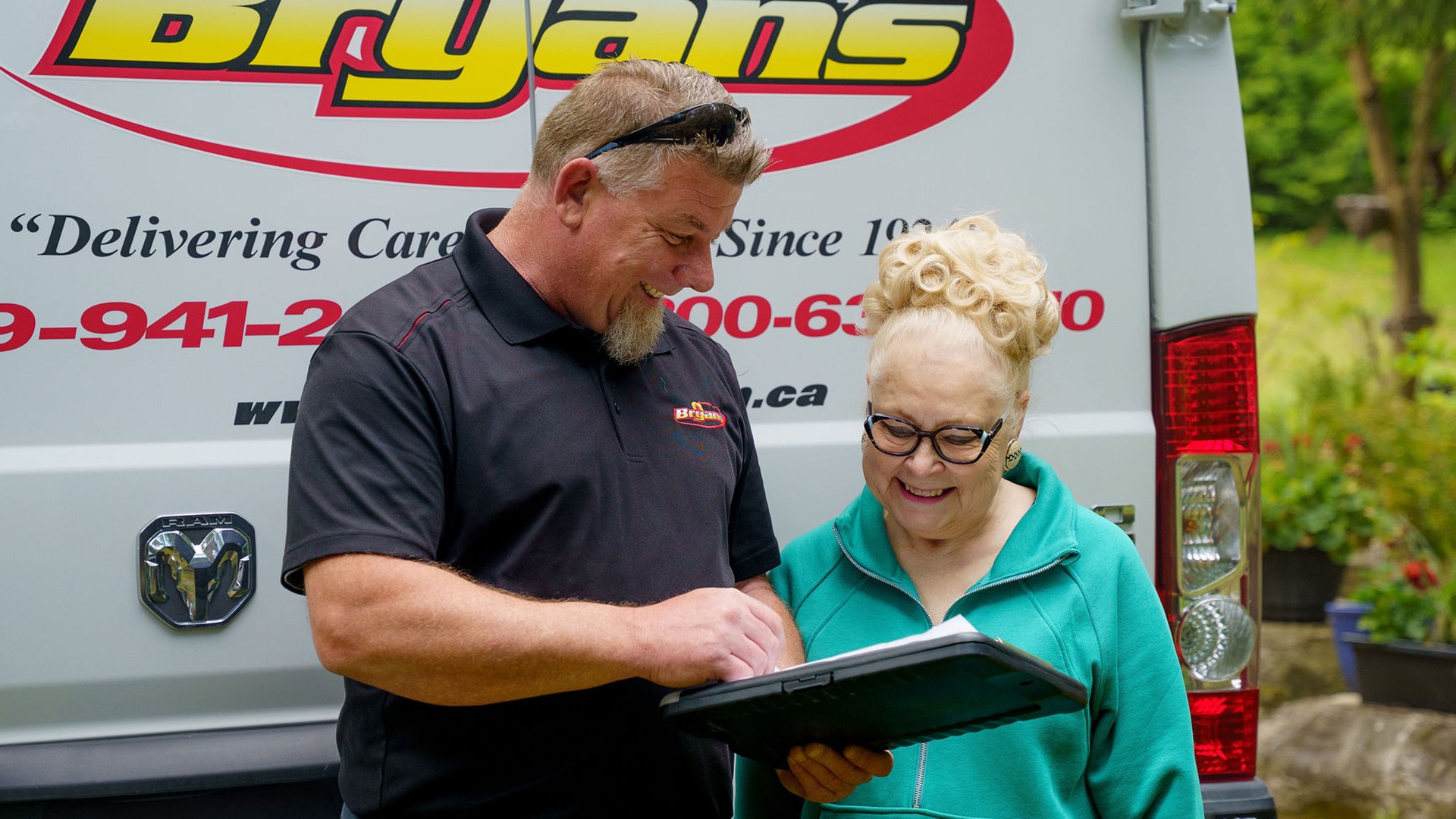 Two people smiling while examining a clipboard in front of a "Bryans" van with a logo. Appears friendly and informal outdoors.