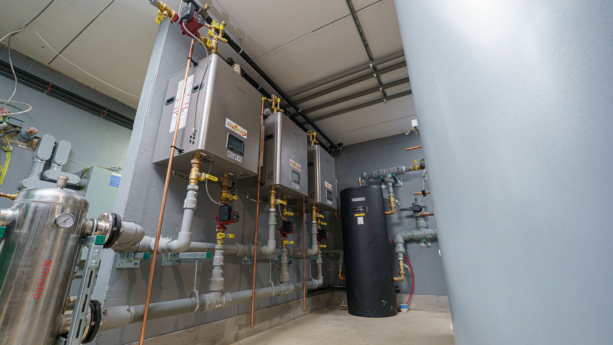 Industrial room with multiple boilers and pipes against a gray wall, featuring a black cylindrical tank and various valves.