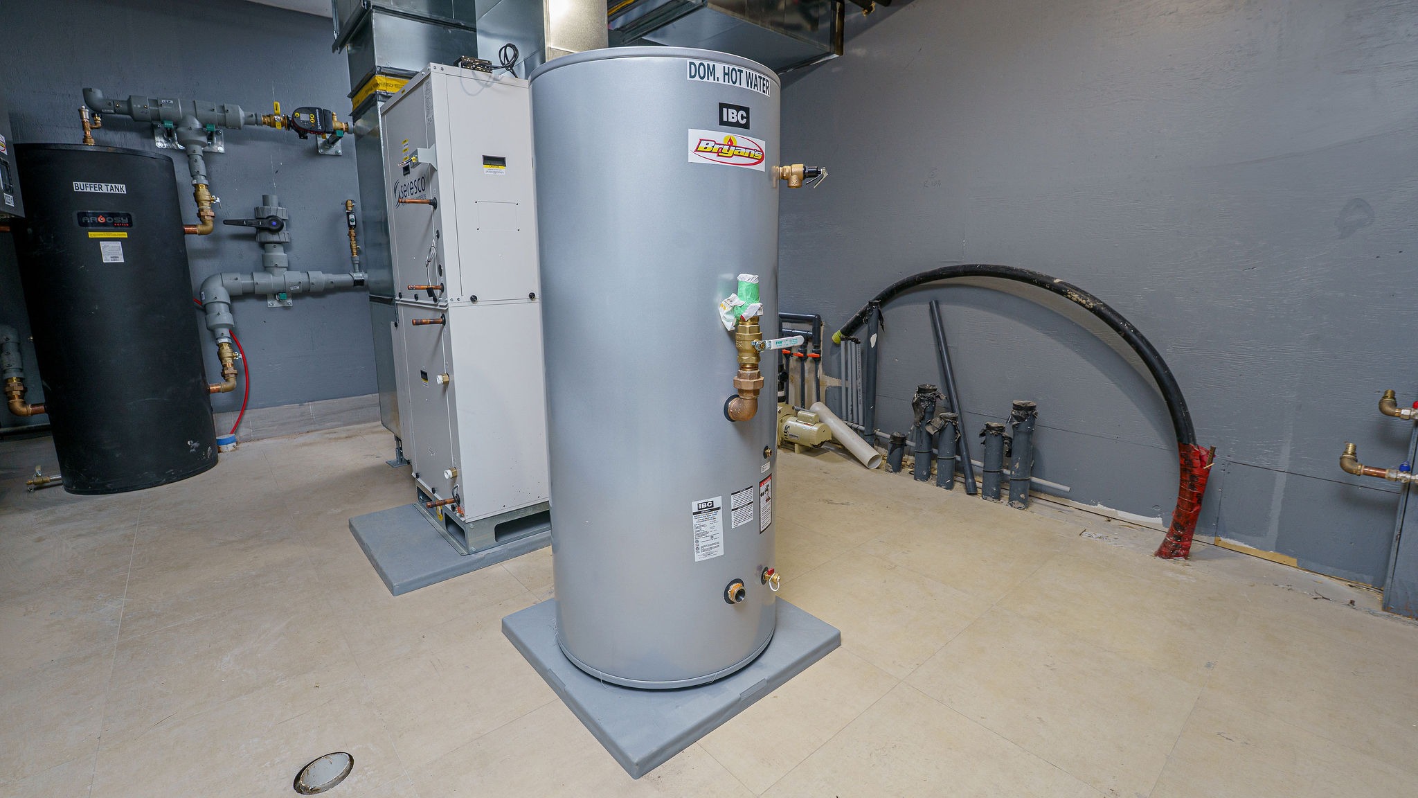 A utility room with a large hot water tank, buffer tank, and complex piping system. Walls and floor are industrial style.