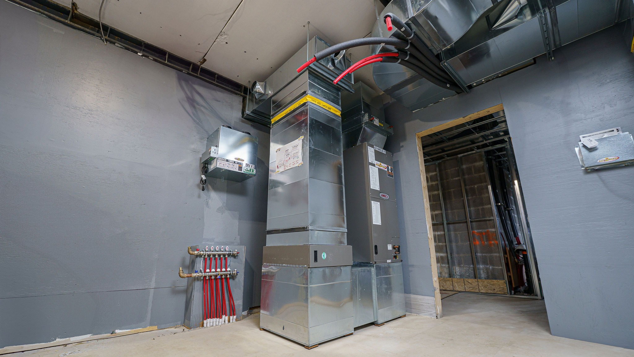 A commercial utility room with large HVAC systems, exposed ductwork, and control panels on a plain grey wall, with pipes leading into the floor.