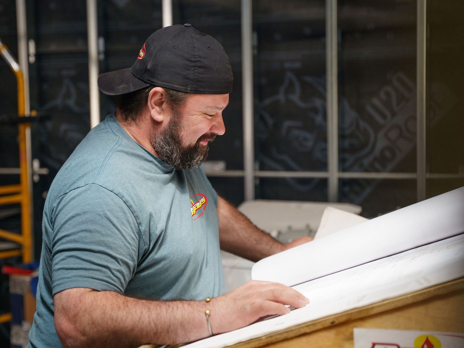 A person examines large blueprint papers indoors, wearing a cap and t-shirt. The setting has construction elements and dim lighting.