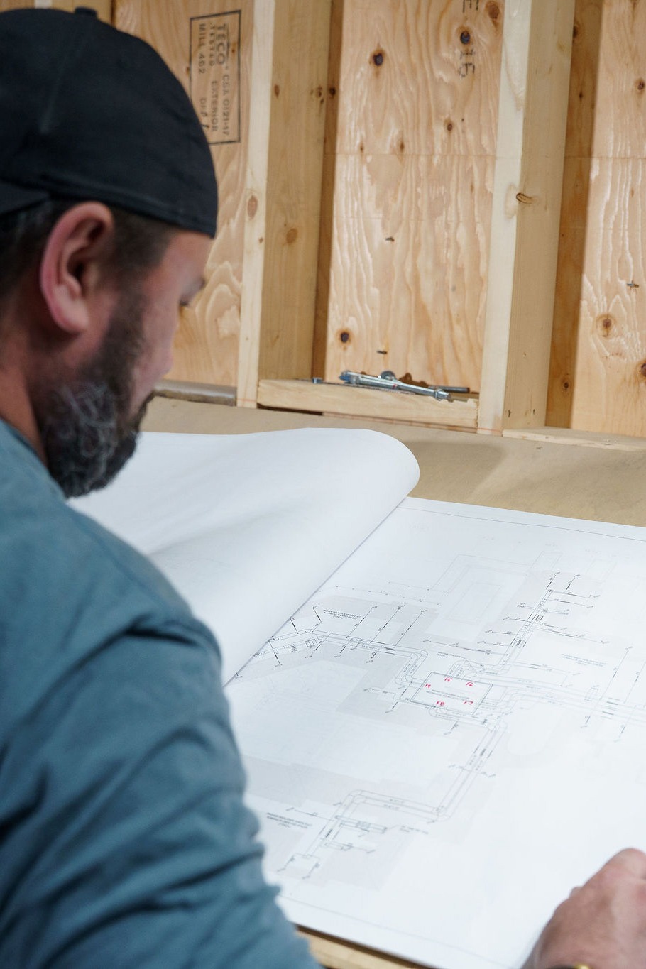A person examines architectural blueprints on a wooden table, set against an unfinished background with exposed wooden panels.