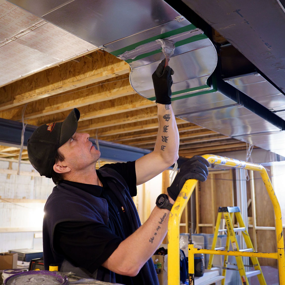 A person on a ladder works on ductwork in a construction site, applying sealant with a putty knife, wearing gloves and a cap.