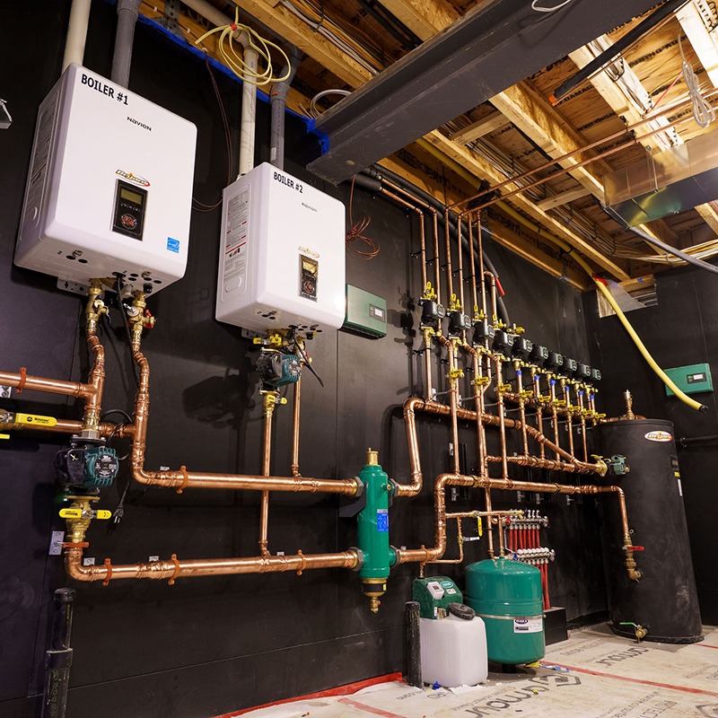 A mechanical room with two boilers, extensive copper piping, and various control valves visible on a utility panel. Ceiling beams are exposed.