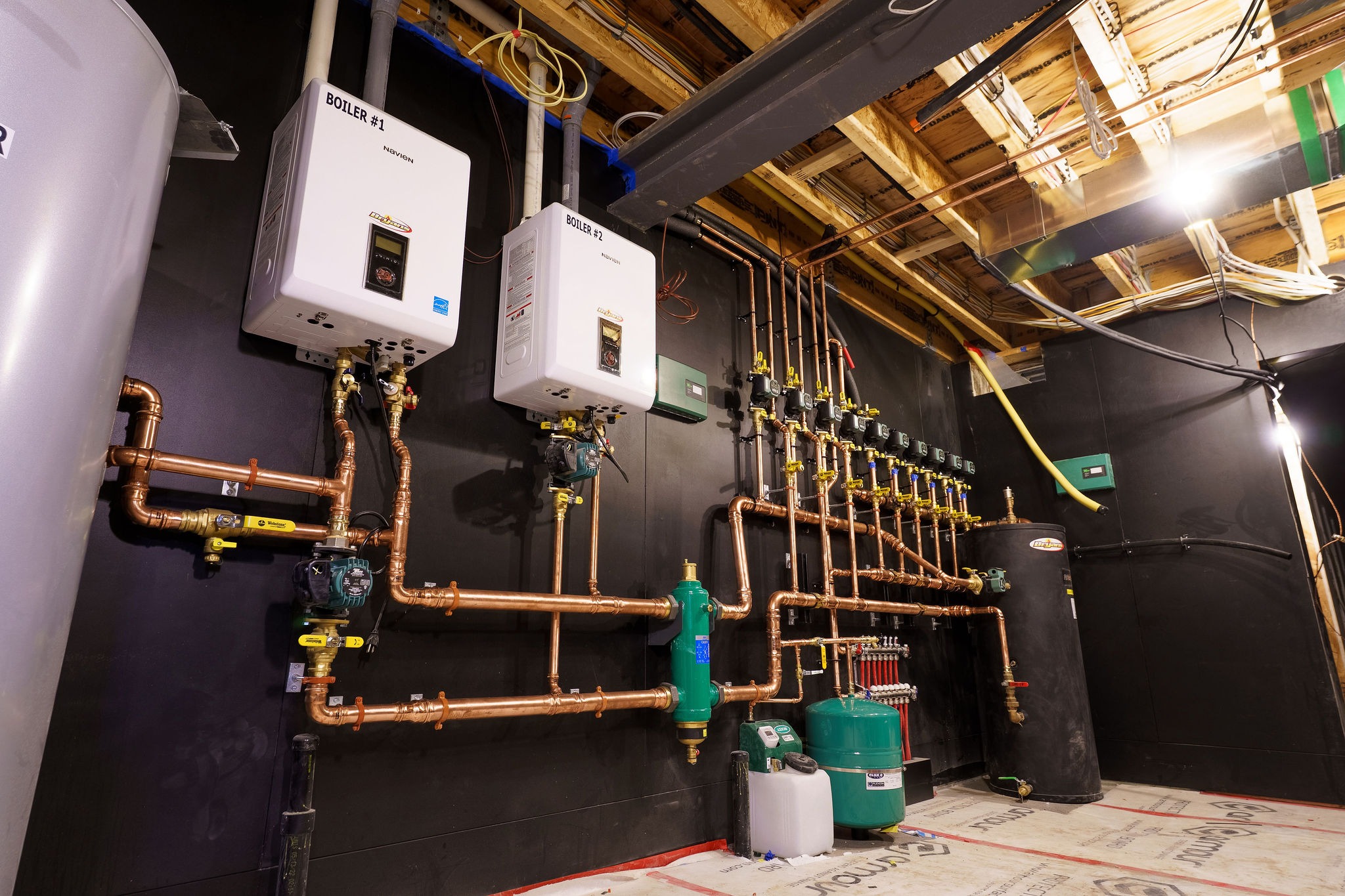 The image shows a utility room with boilers, copper pipes, and a water tank, illuminated by ceiling lights.