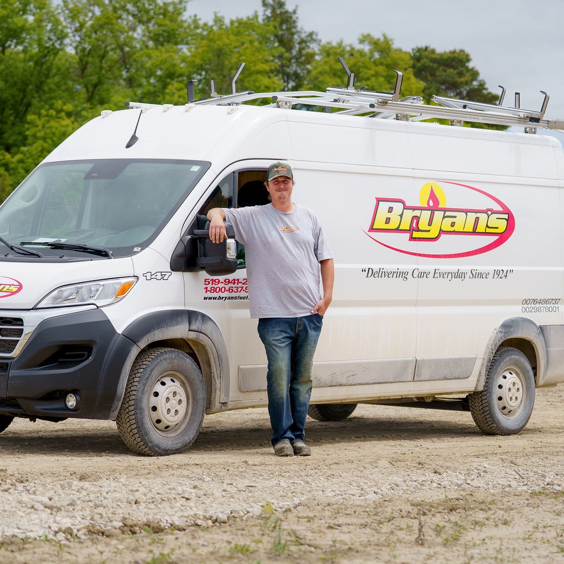 A person leans against a white work van with "Bryan's" branding, surrounded by trees and dirt ground, conveying a casual outdoor scene.