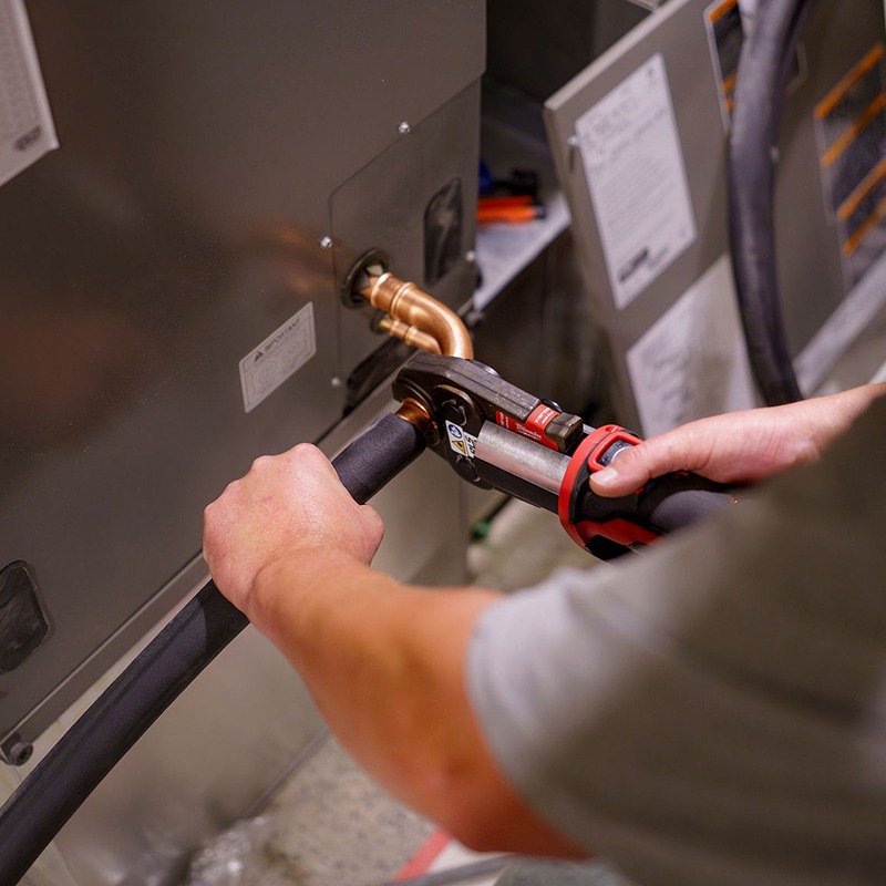 A person uses a tool to connect a hose to HVAC equipment, working on maintenance or installation in a mechanical room.
