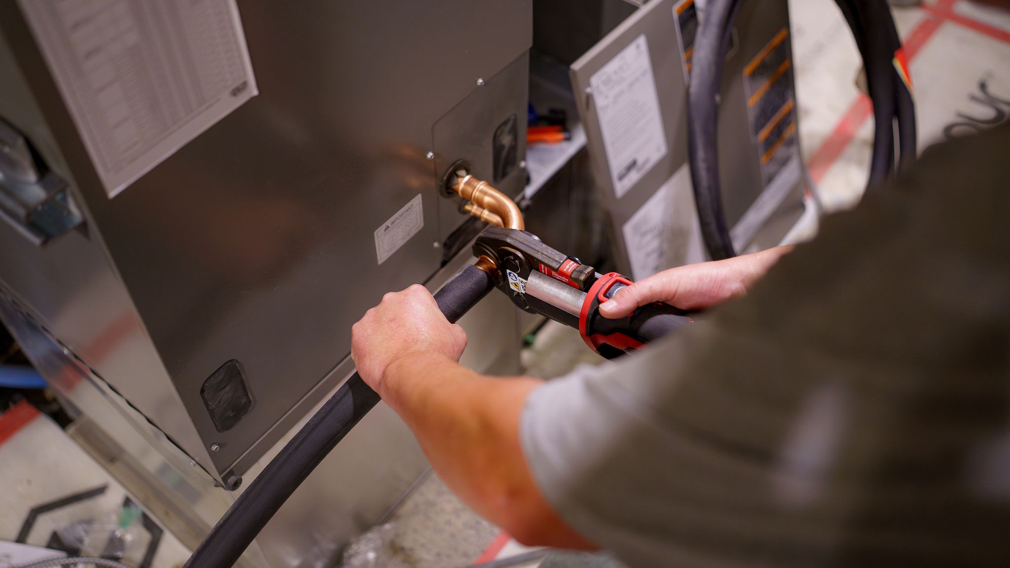 A person uses a hydraulic tool to connect insulated tubing to a metal unit, likely part of an HVAC system installation process.