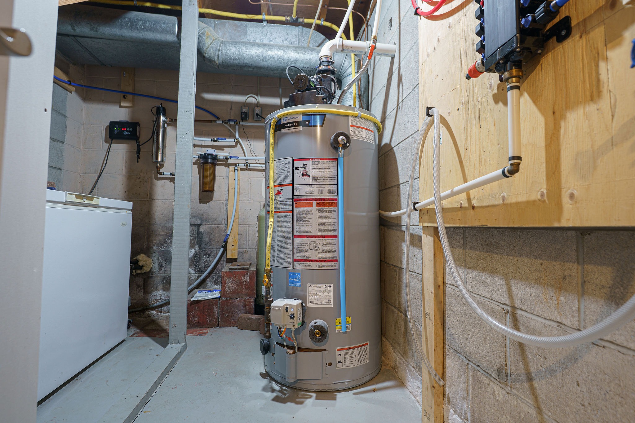 A basement features a water heater, white appliance, and piping on concrete walls, surrounded by various utility connections and cables.