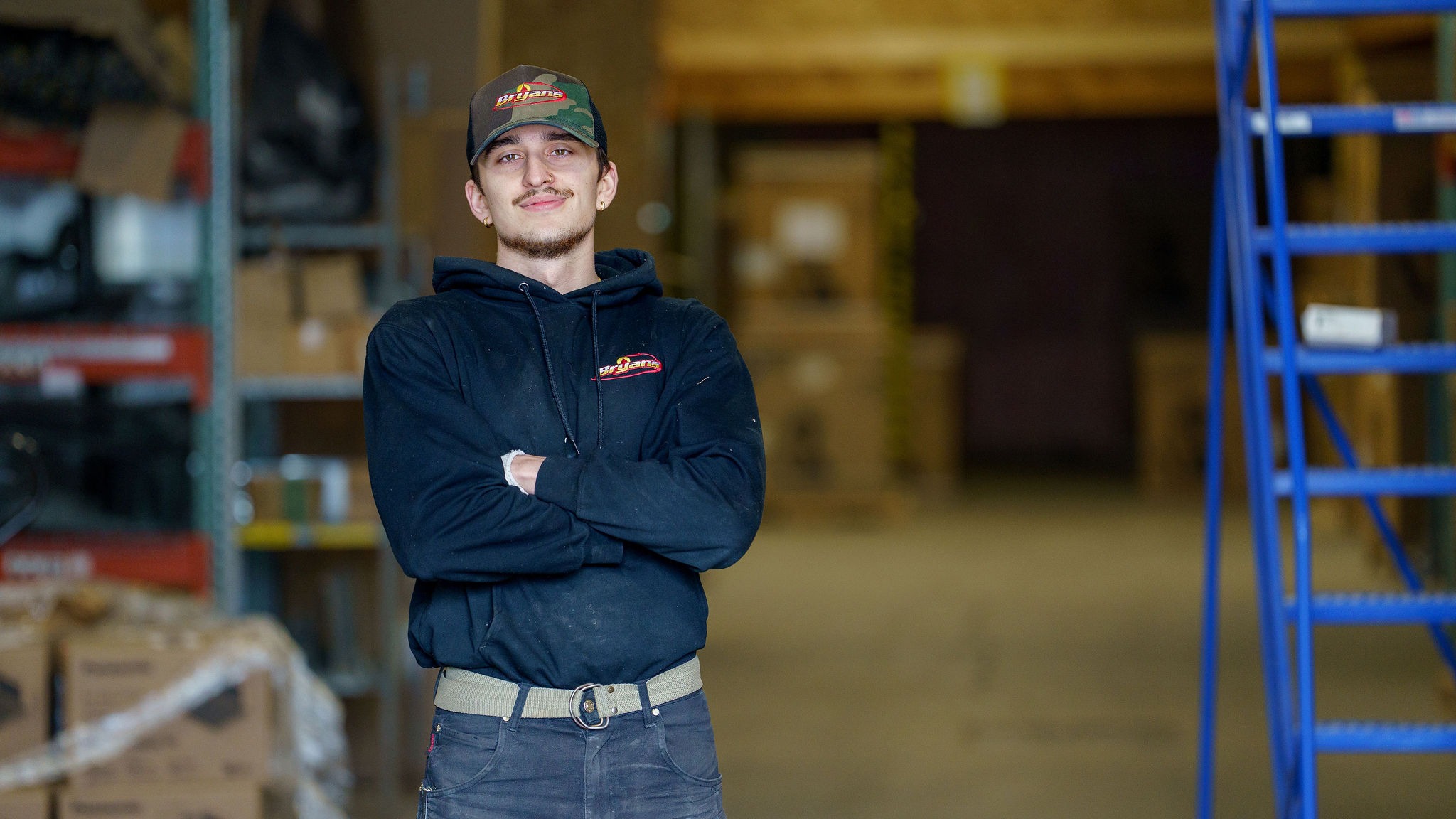 A person in a black hoodie and cap stands confidently in a warehouse setting, with shelves and boxes in the background.