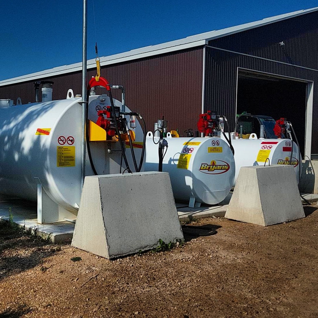 Outdoor scene with large white diesel fuel tanks beside a metal building. Blue sky above. Concrete barriers in the foreground.