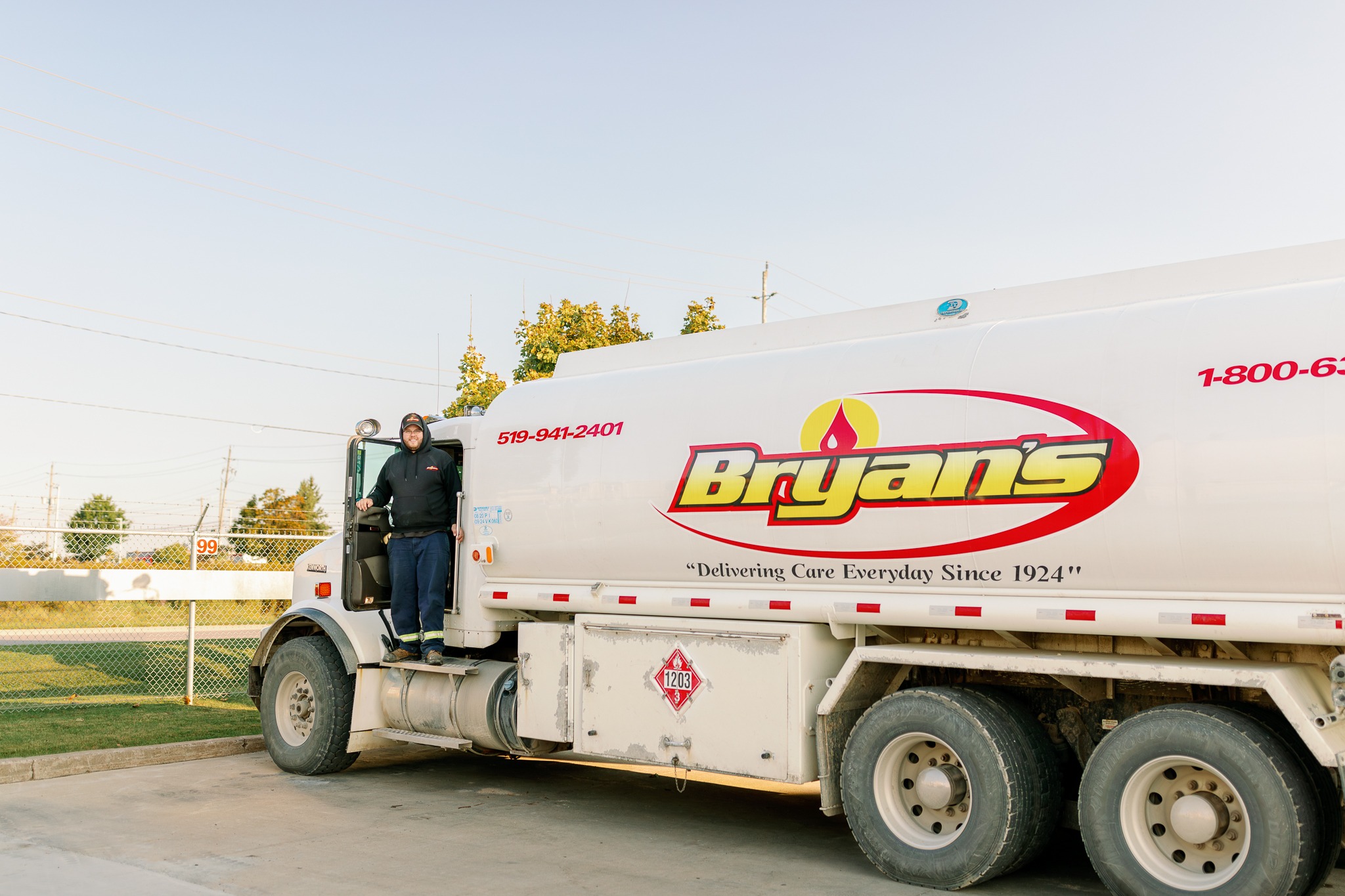 A person stands beside a large fuel truck labeled "Bryan's" in a fenced outdoor area under a clear blue sky.