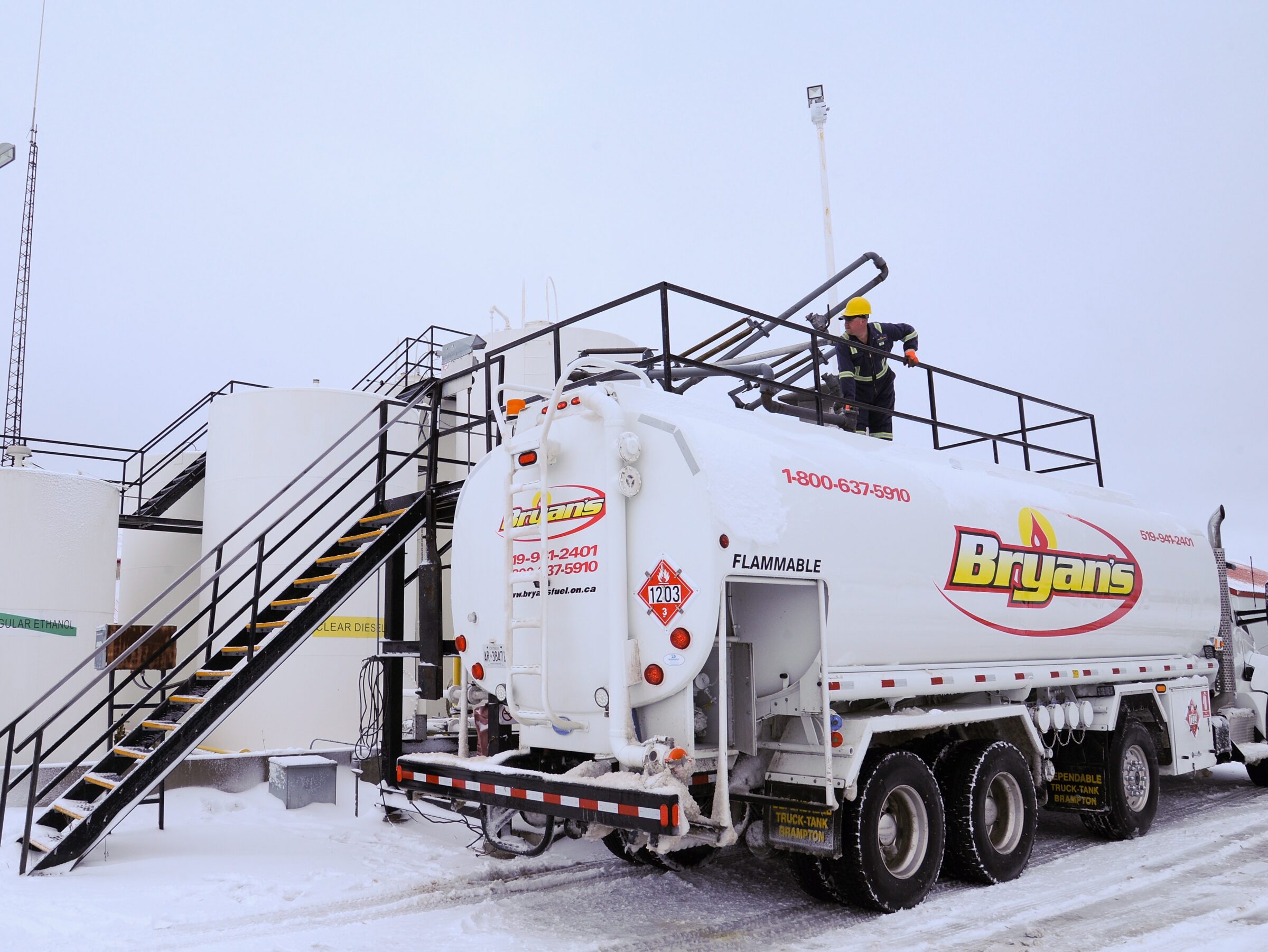 A person in safety gear manages a Bryans fuel truck beside industrial tanks, set on a snowy ground, with a sky overcast.
