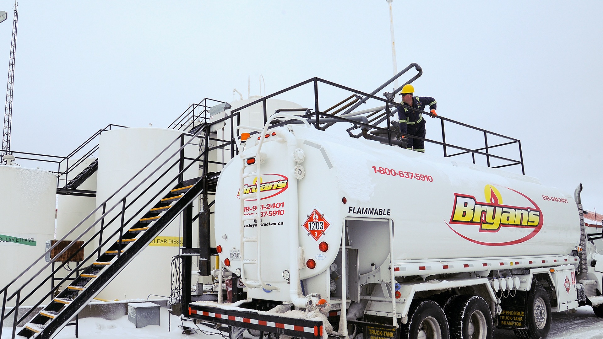 Person performing maintenance on a fuel truck by large white storage tanks covered in snow, labeled "Bryan's," amid industrial surroundings.