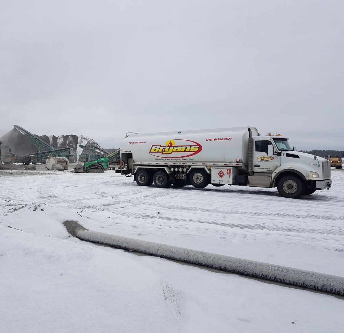 A Bryan's fuel truck is parked on a snowy industrial site with machinery and large rock piles in the background.