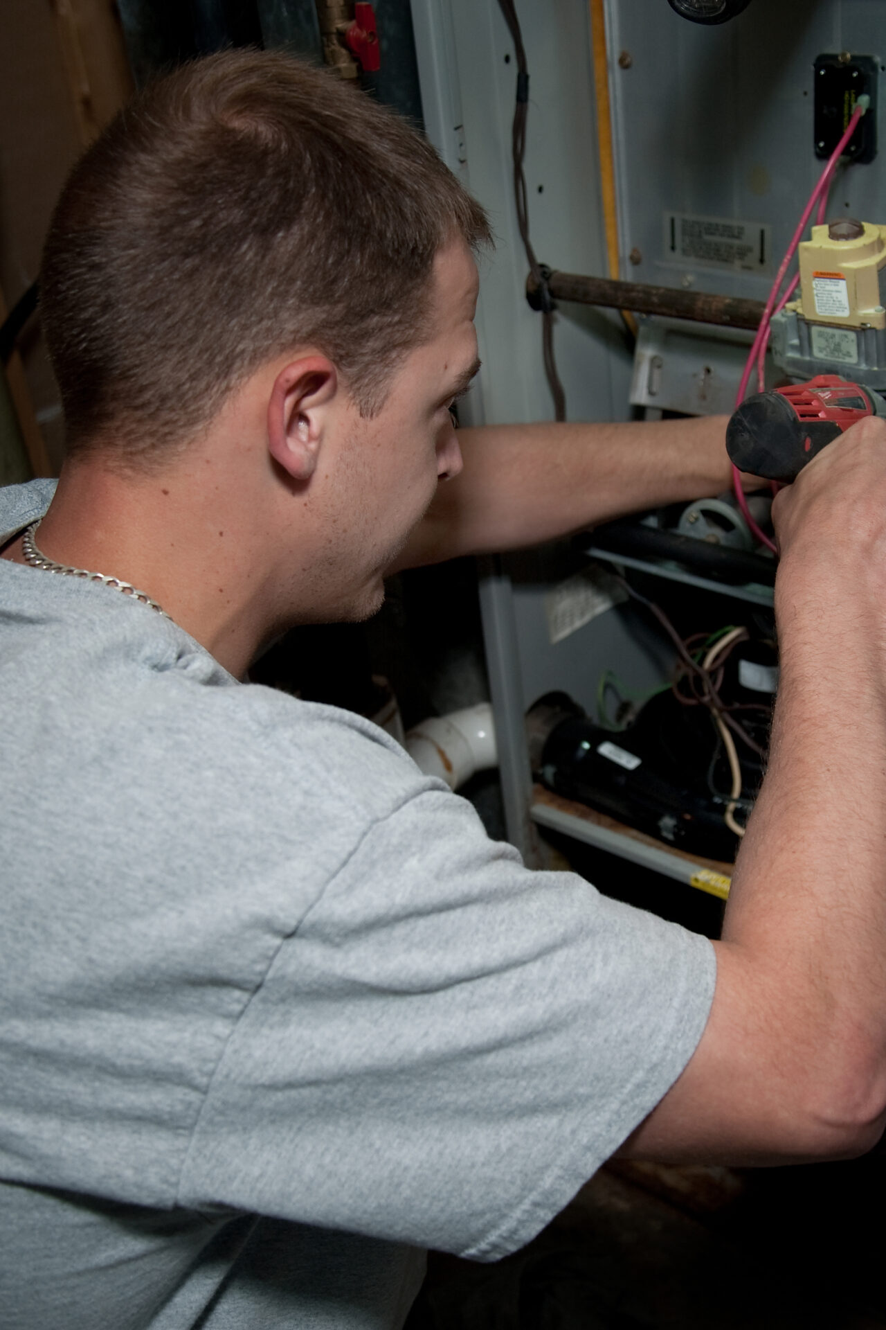 A person in a gray shirt is using a drill to work on wiring inside an open utility panel.