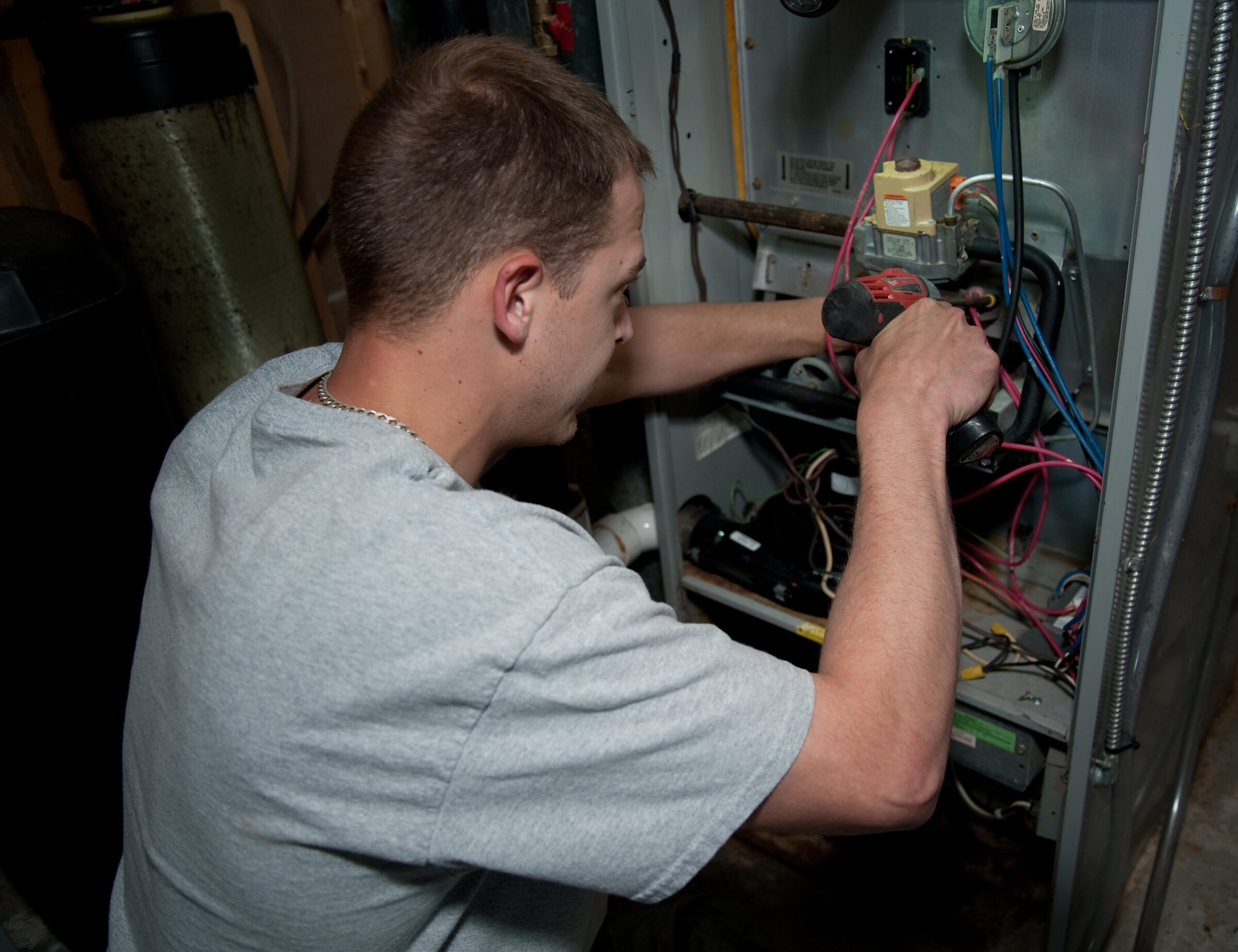 A person in a gray shirt repairs an appliance, using a drill and adjusting wires in an industrial setting.