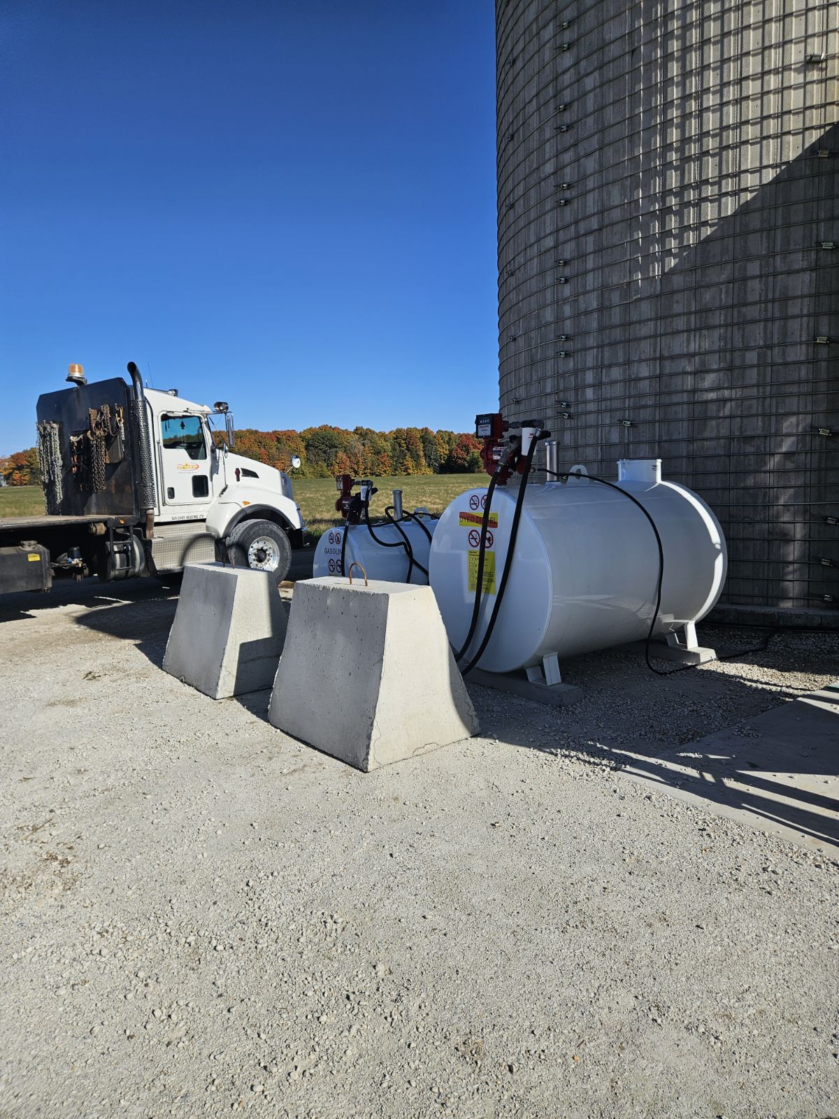A truck parked next to cylindrical tanks and concrete barriers under clear blue sky, with autumnal trees in the background.