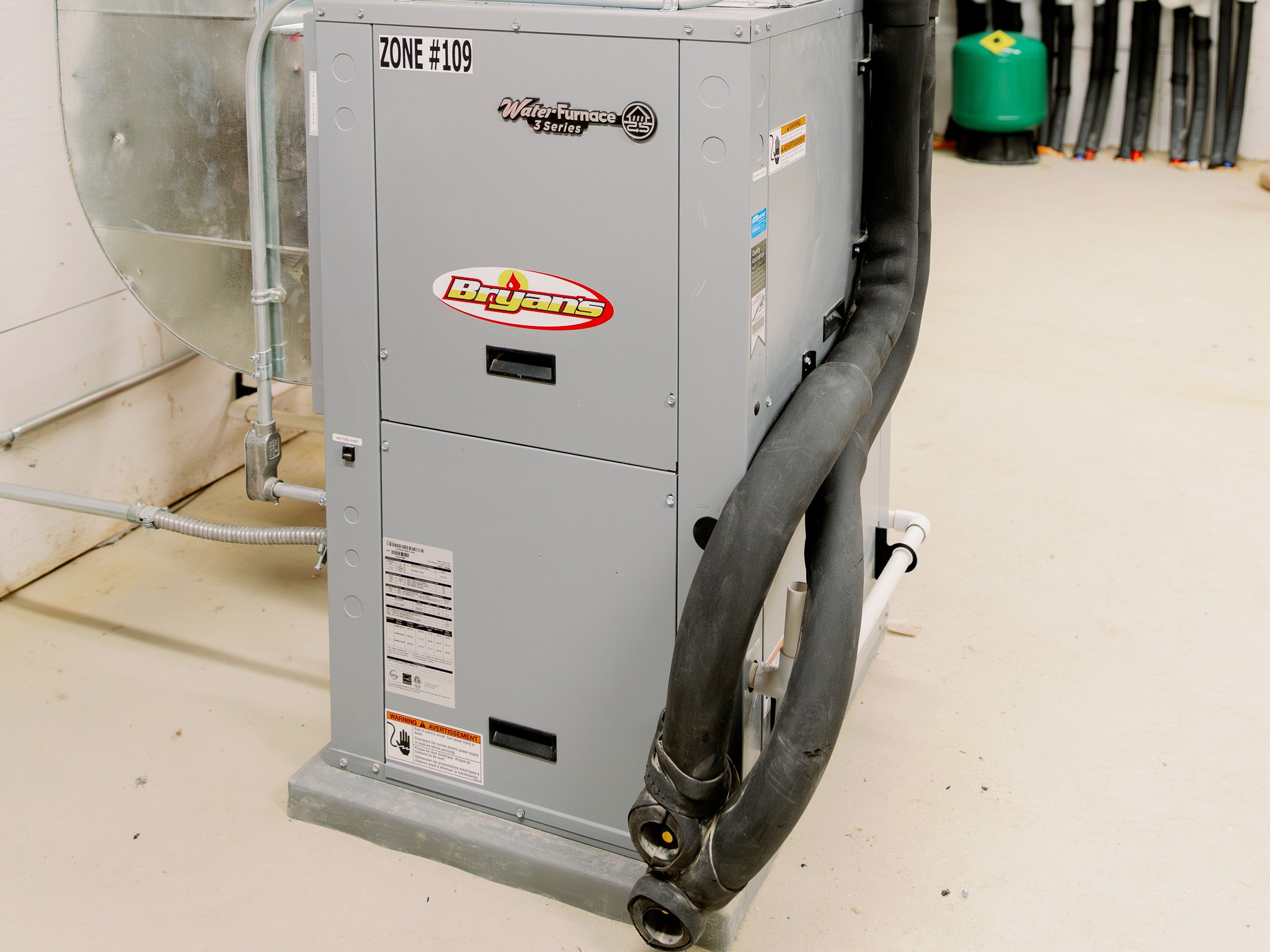 A geothermal heating system unit is installed indoors. It features gray metal casing, black pipes, and multiple labels, set on a concrete floor.