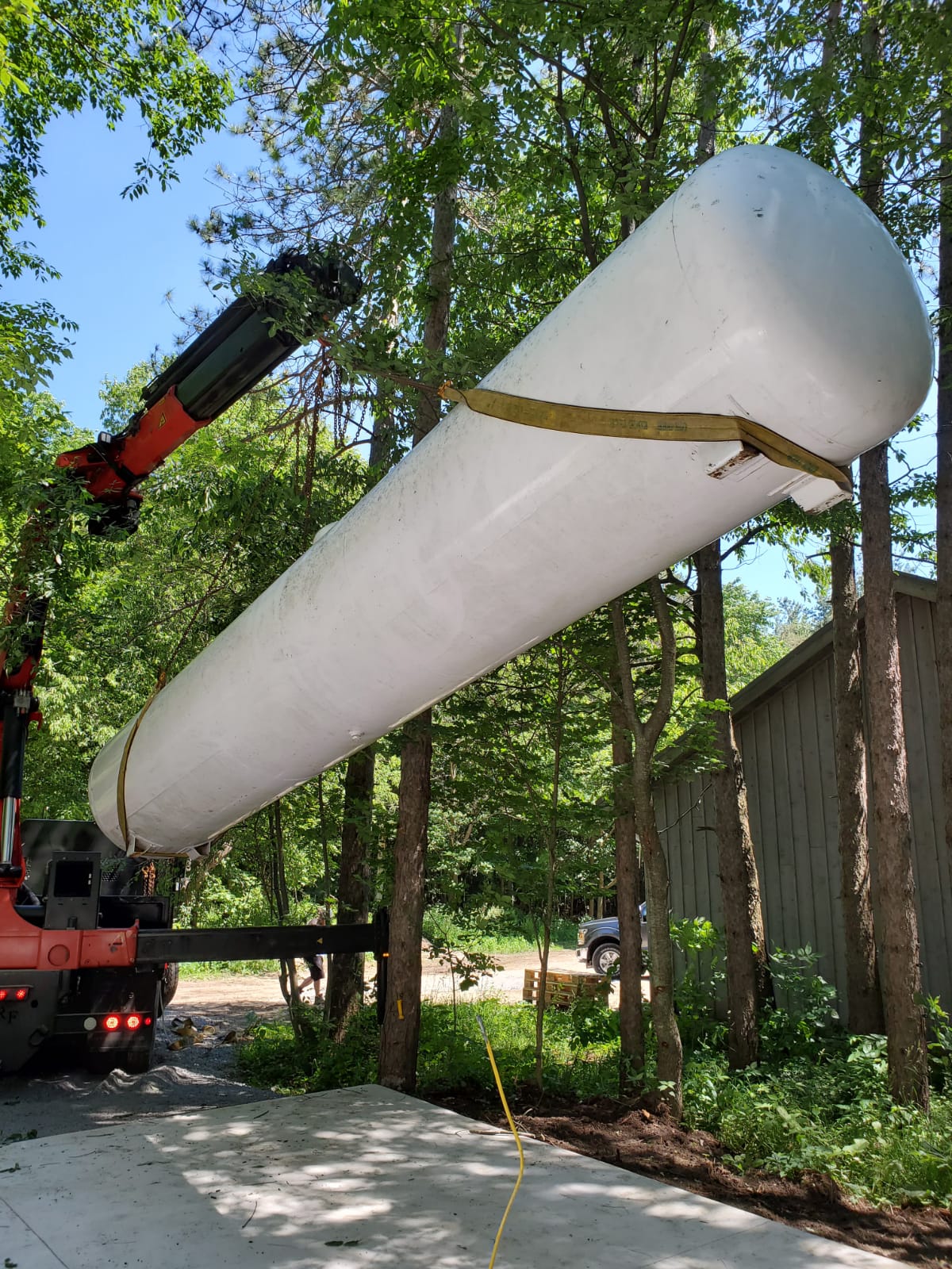 A crane lifting a large, white tank among trees and a building, with a truck visible in the background under clear skies.