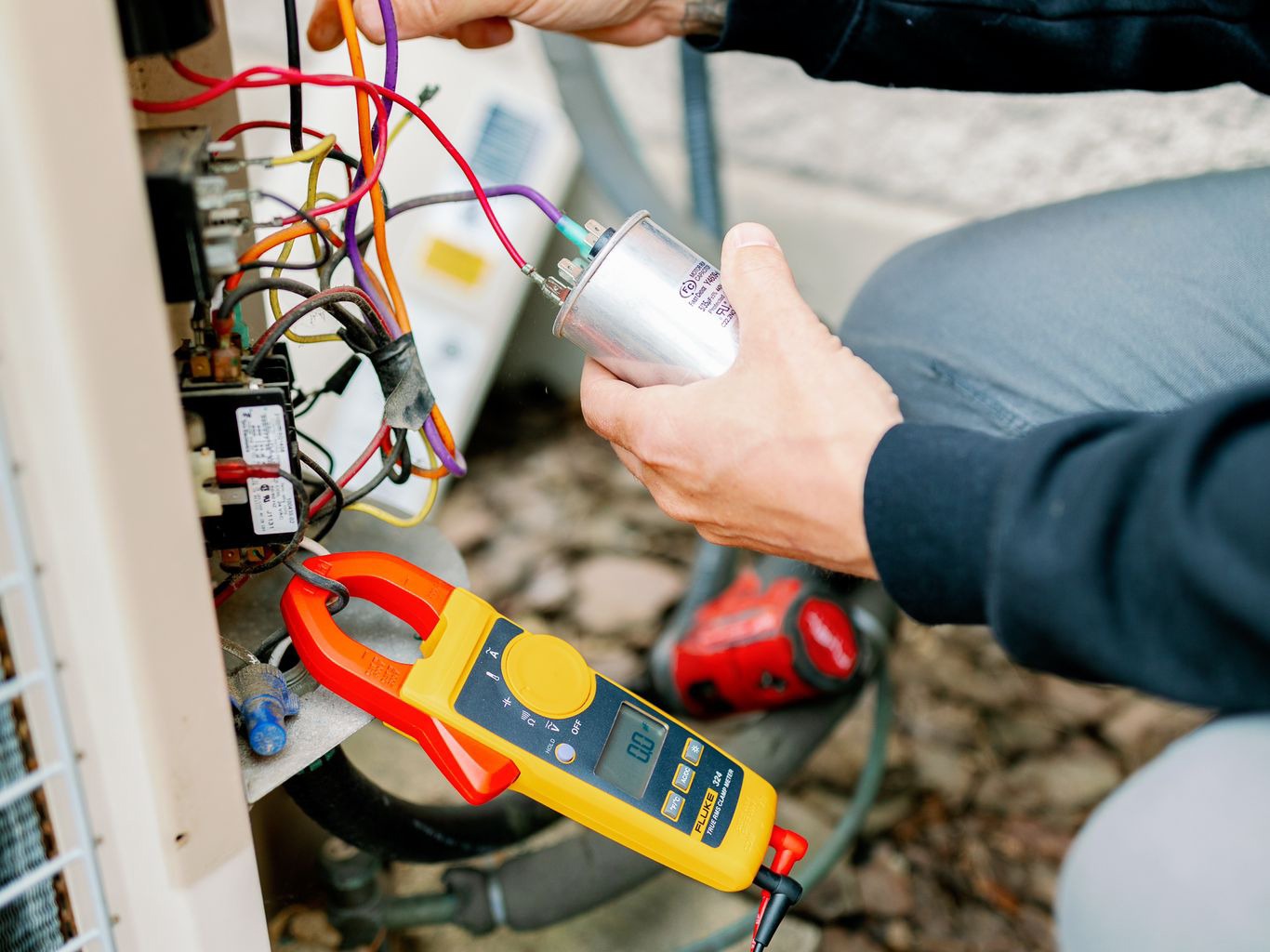 A person repairs an electrical unit outdoors, holding a capacitor and using a digital multimeter for diagnostics.