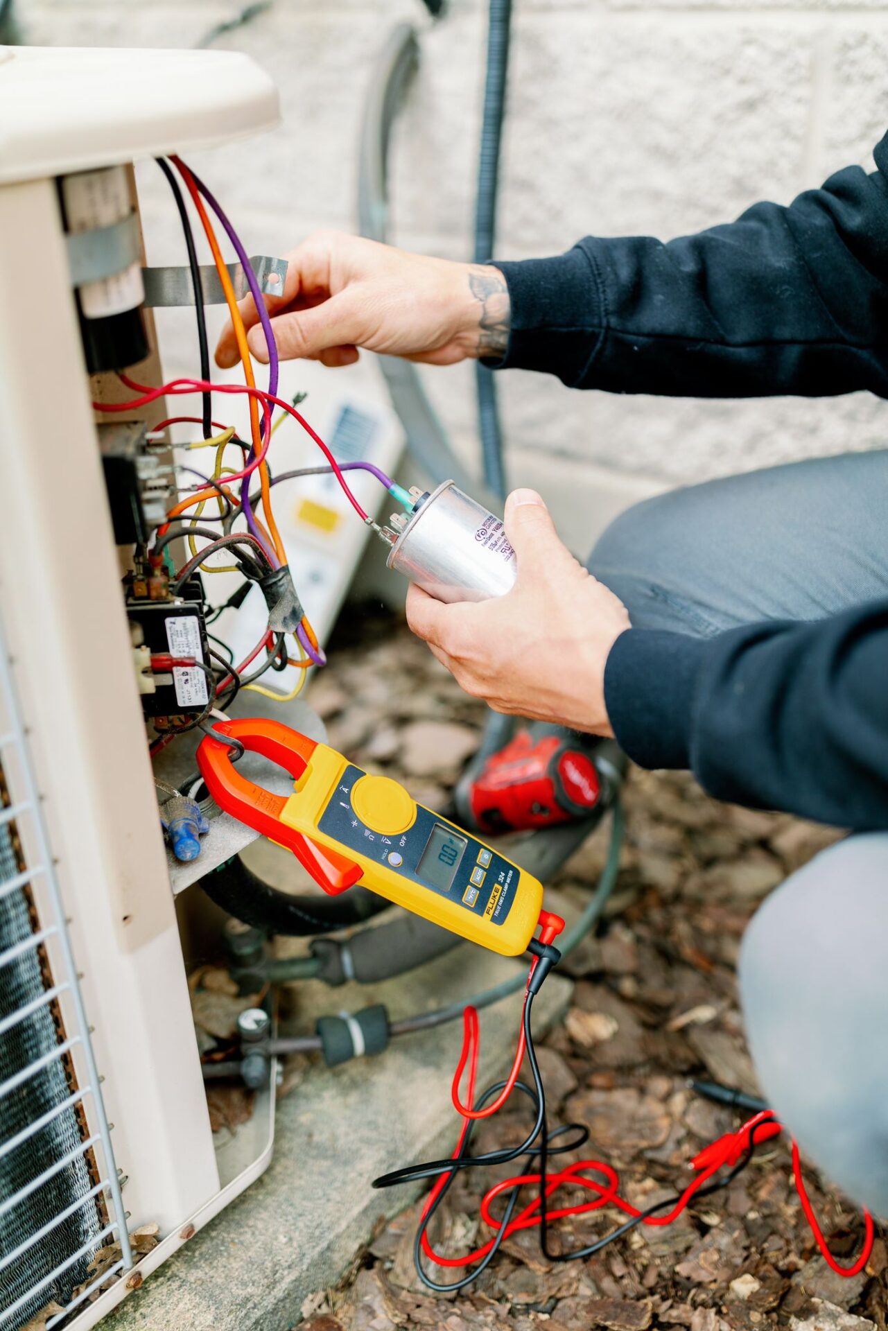 A person inspects an HVAC unit, using tools and colorful wires. A multimeter is attached, amidst an outdoor environment with scattered leaves.