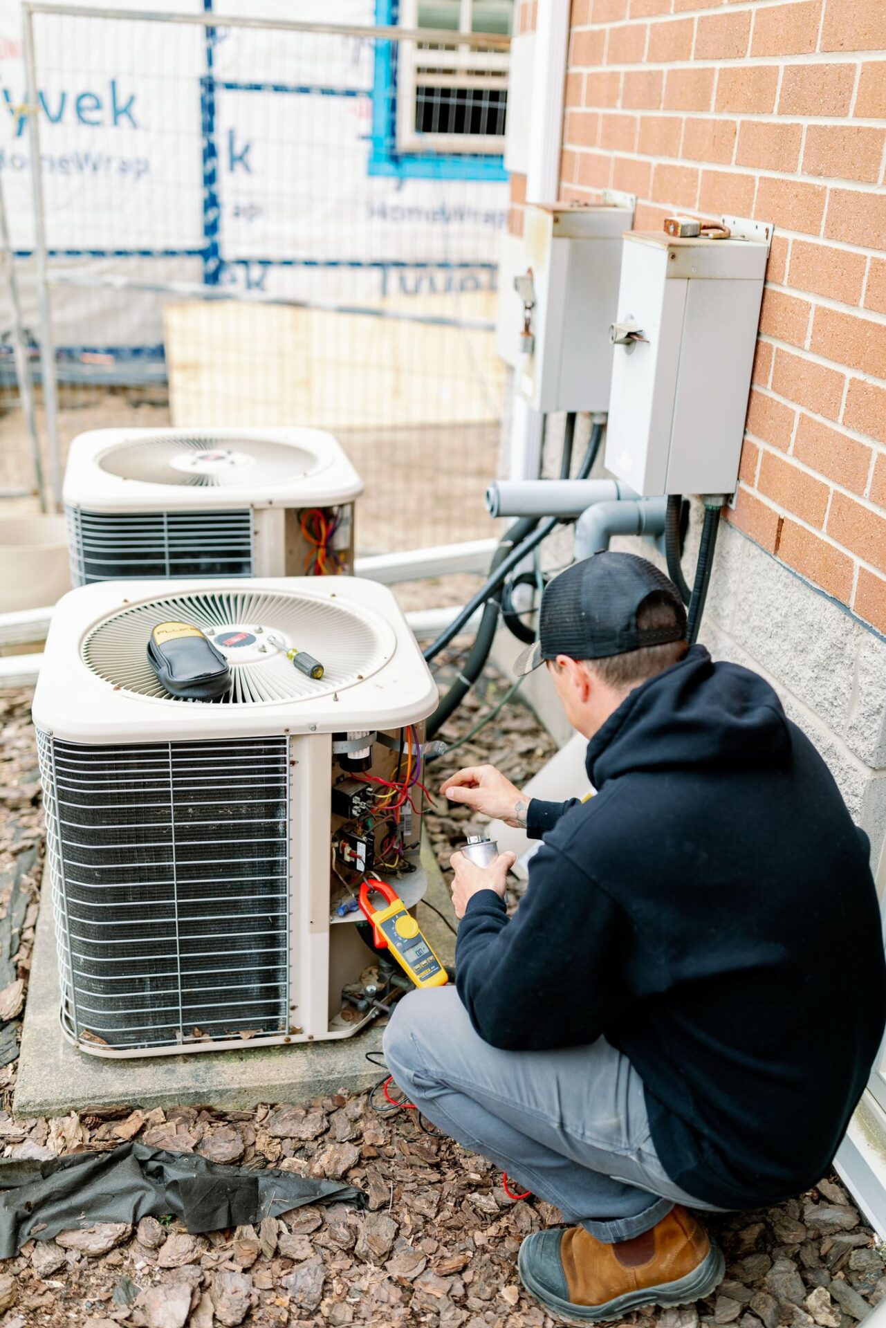 A person in a cap repairs an air conditioning unit outside a brick building under construction, with tools and electrical wiring visible.