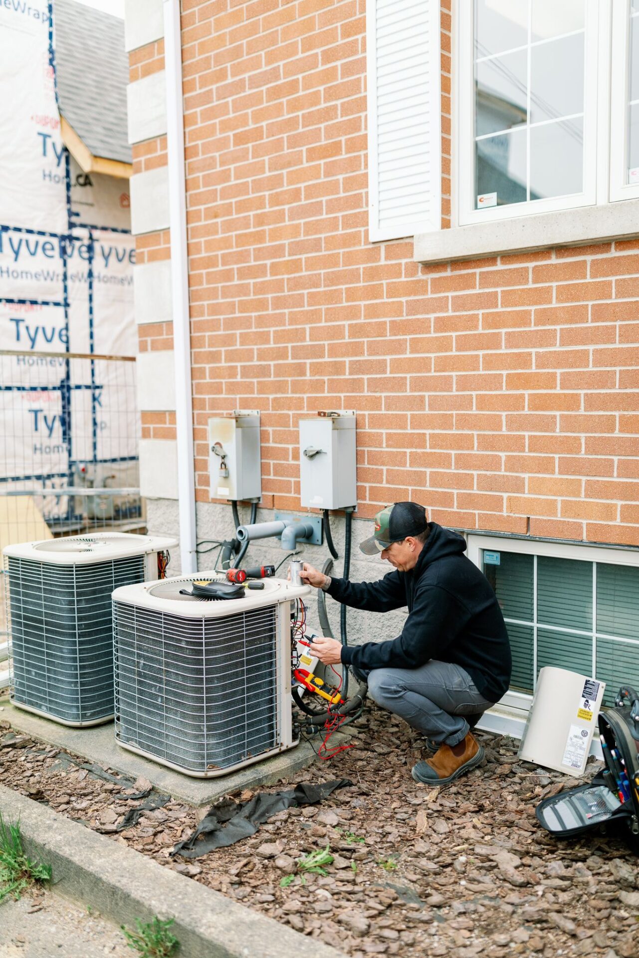 A person in a black hoodie is repairing an HVAC unit outside a brick house, with tools and equipment nearby.
