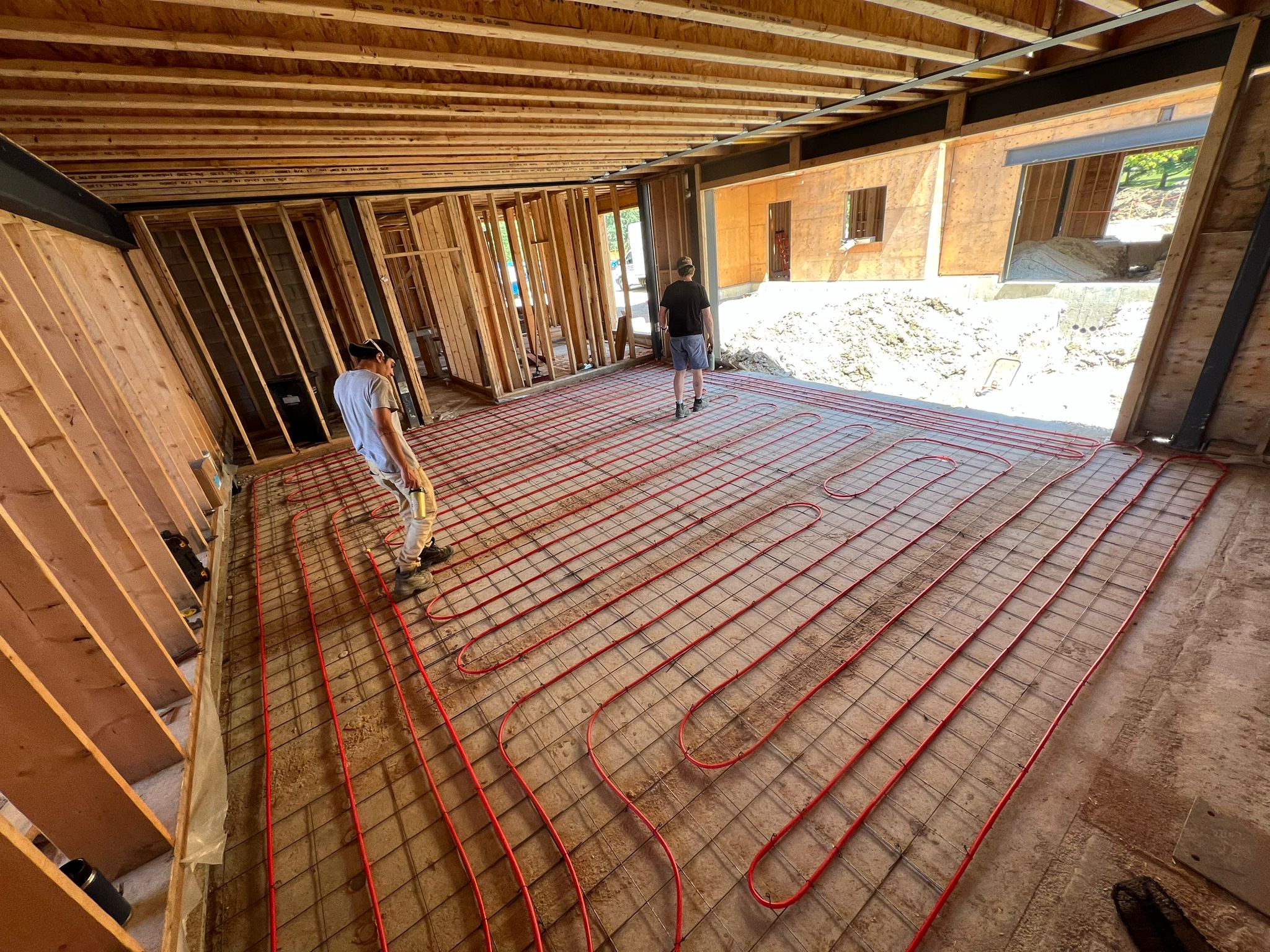 Two people work on installing radiant floor heating in a wooden-framed building under construction. No distinguishable landmarks are present.
