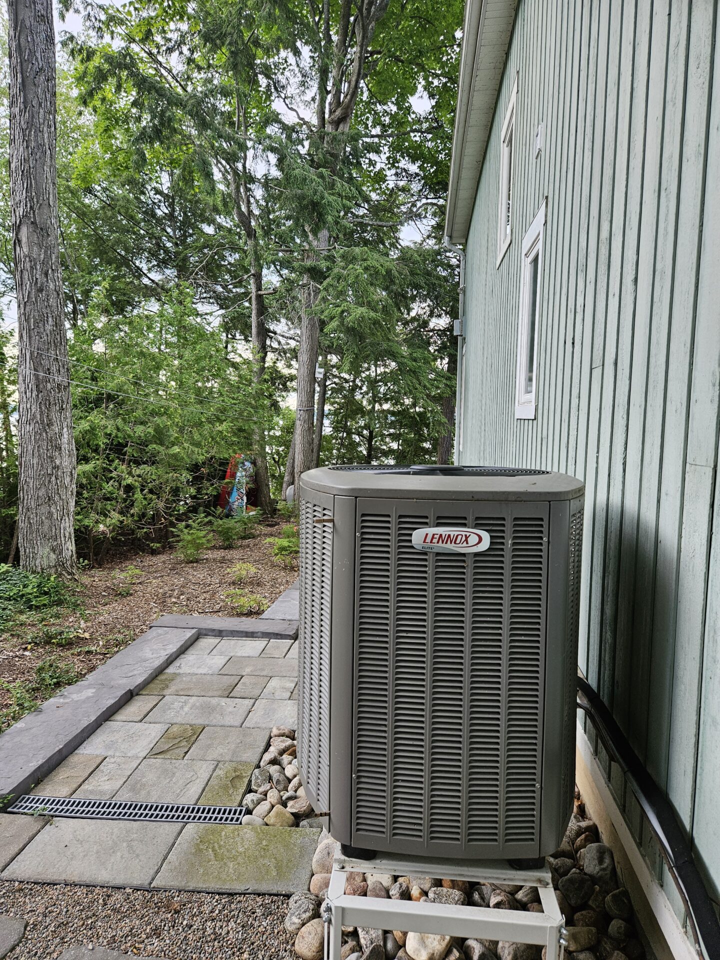 An outdoor air conditioning unit beside a wooden house, surrounded by trees and a stone path, on a cloudy day.