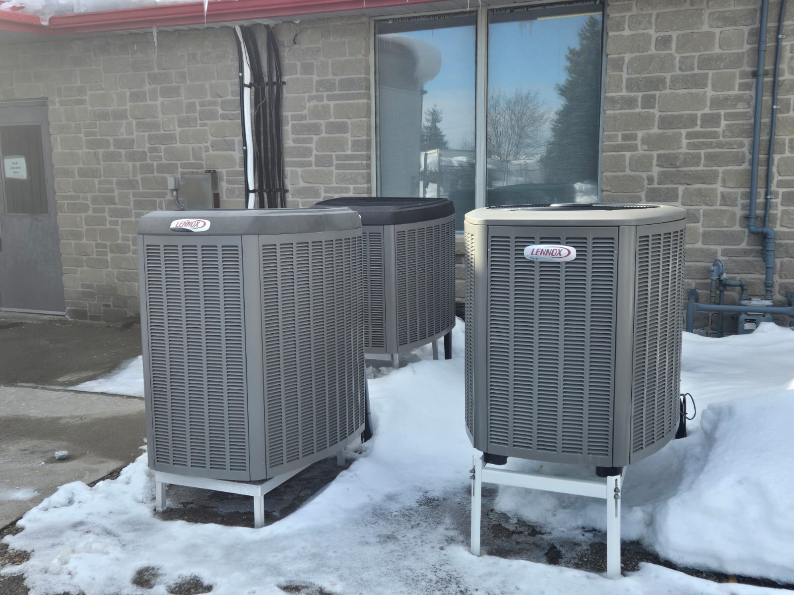 Three Lennox air conditioning units are outside a brick building surrounded by snow, under a clear blue sky with visible trees in the reflection.