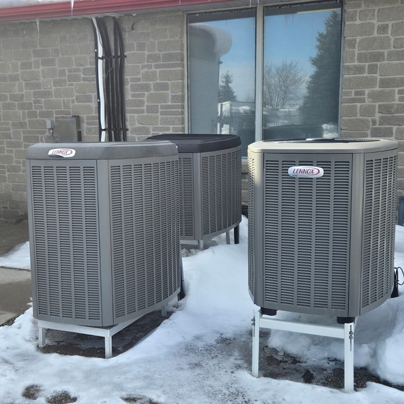Three outdoor Lennox air conditioning units stand on snow-covered ground near a brick building with windows reflecting a clear sky.