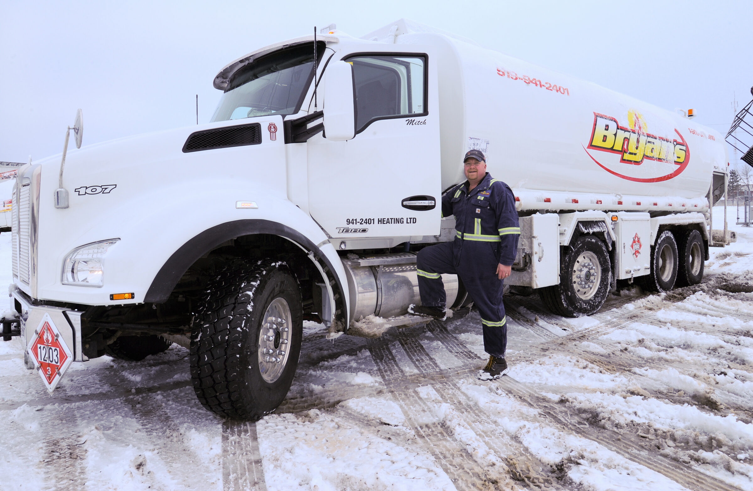 A person in reflective gear stands beside a large white fuel truck in a snowy environment. The truck has BryJan's branding.