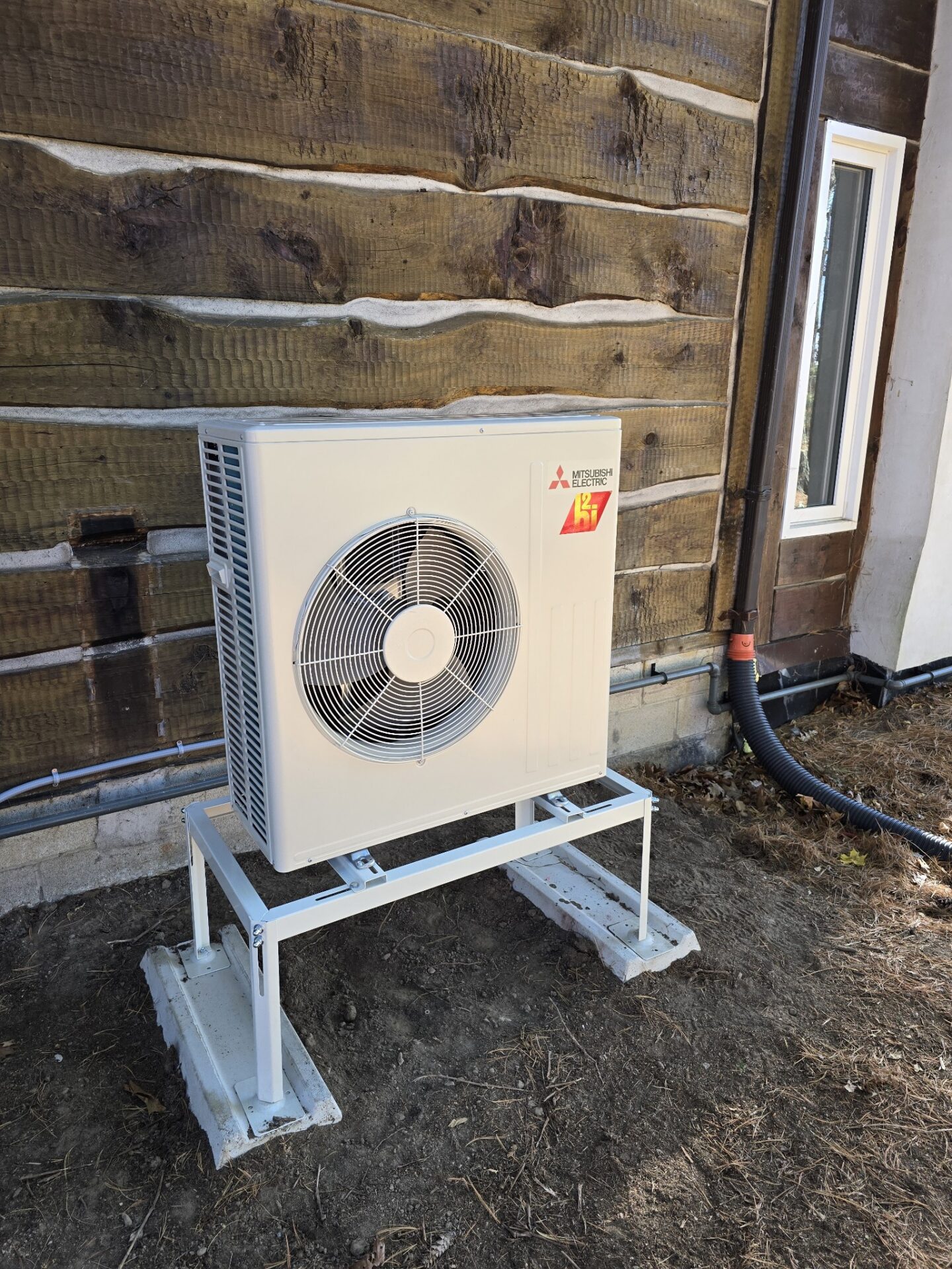 An outdoor Mitsubishi Electric air conditioning unit mounted against a log cabin wall, surrounded by dirt and a small window nearby.