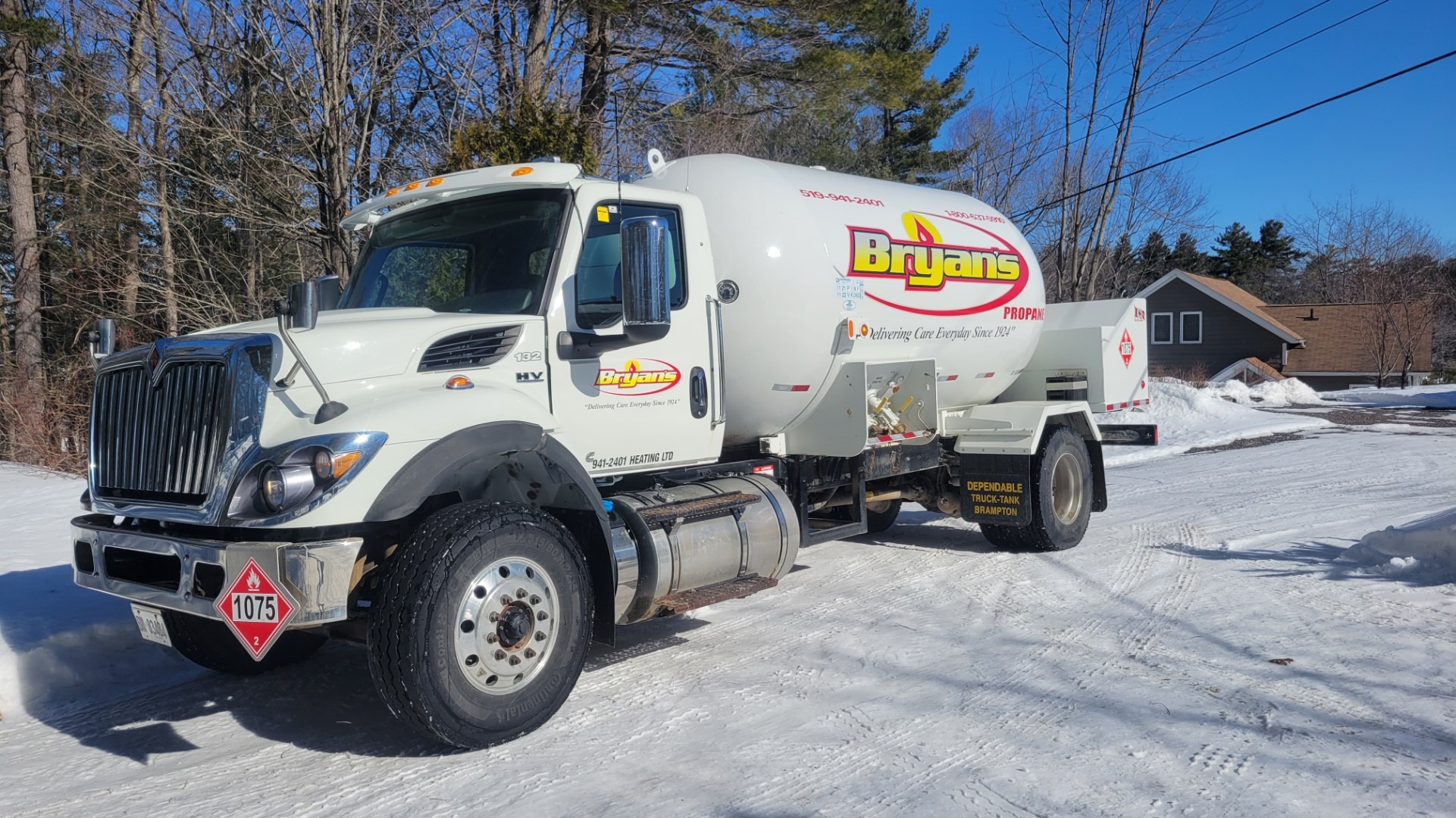 A propane delivery truck parked on a snowy road, surrounded by trees. A house is visible in the background under clear blue skies.
