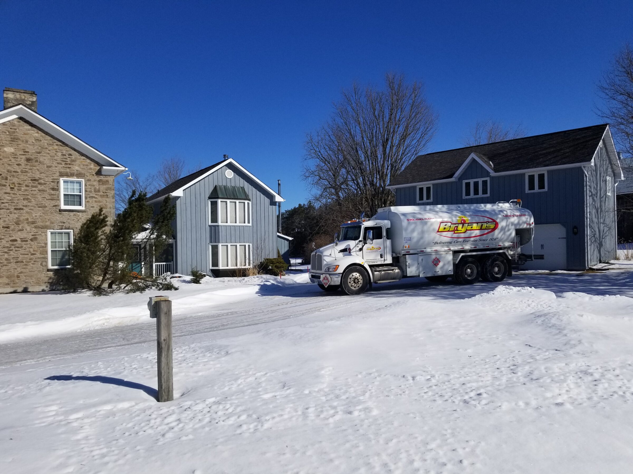 A truck is parked on a snowy street next to two houses, under a clear blue sky, surrounded by bare trees.