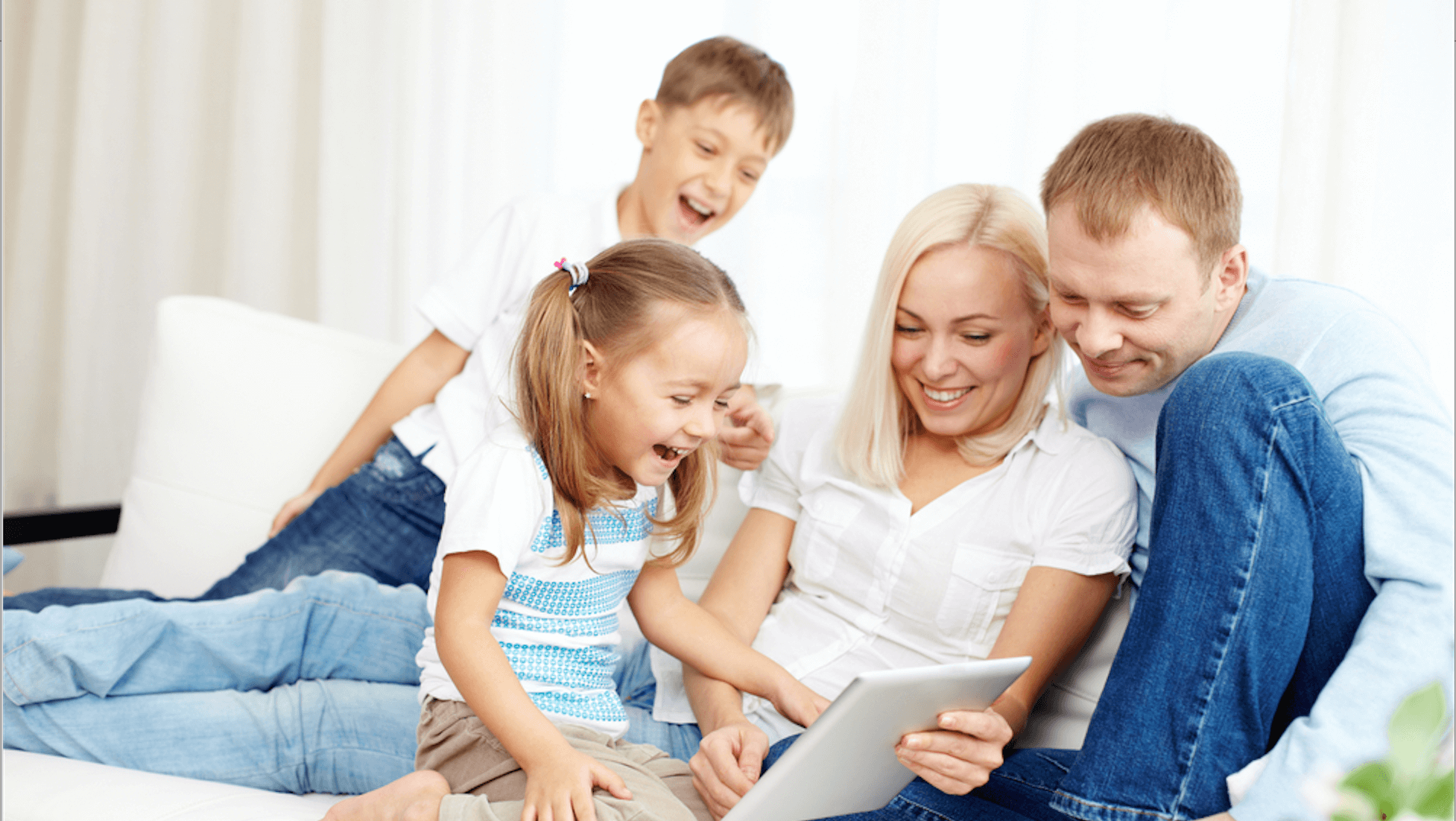A family of four happily gathers around a tablet on a couch in a bright, cozy living room.