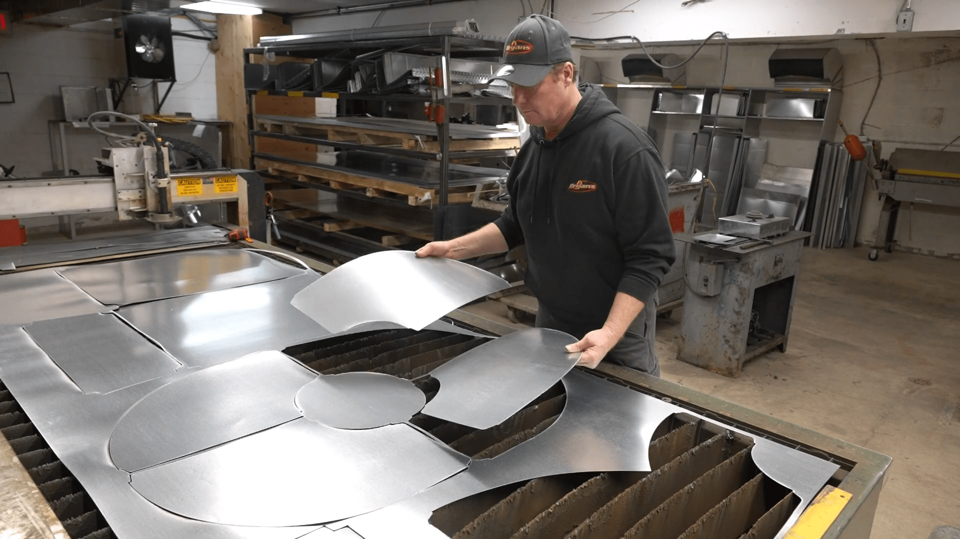 A person in a workshop examines cut sheet metal pieces, with shelves and machinery in the background. The environment suggests industrial work.