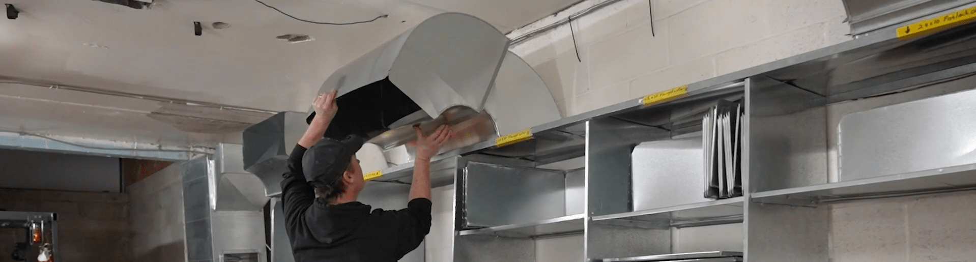 A person installs a large metallic ventilation duct onto a ceiling, working in an industrial or commercial building environment.